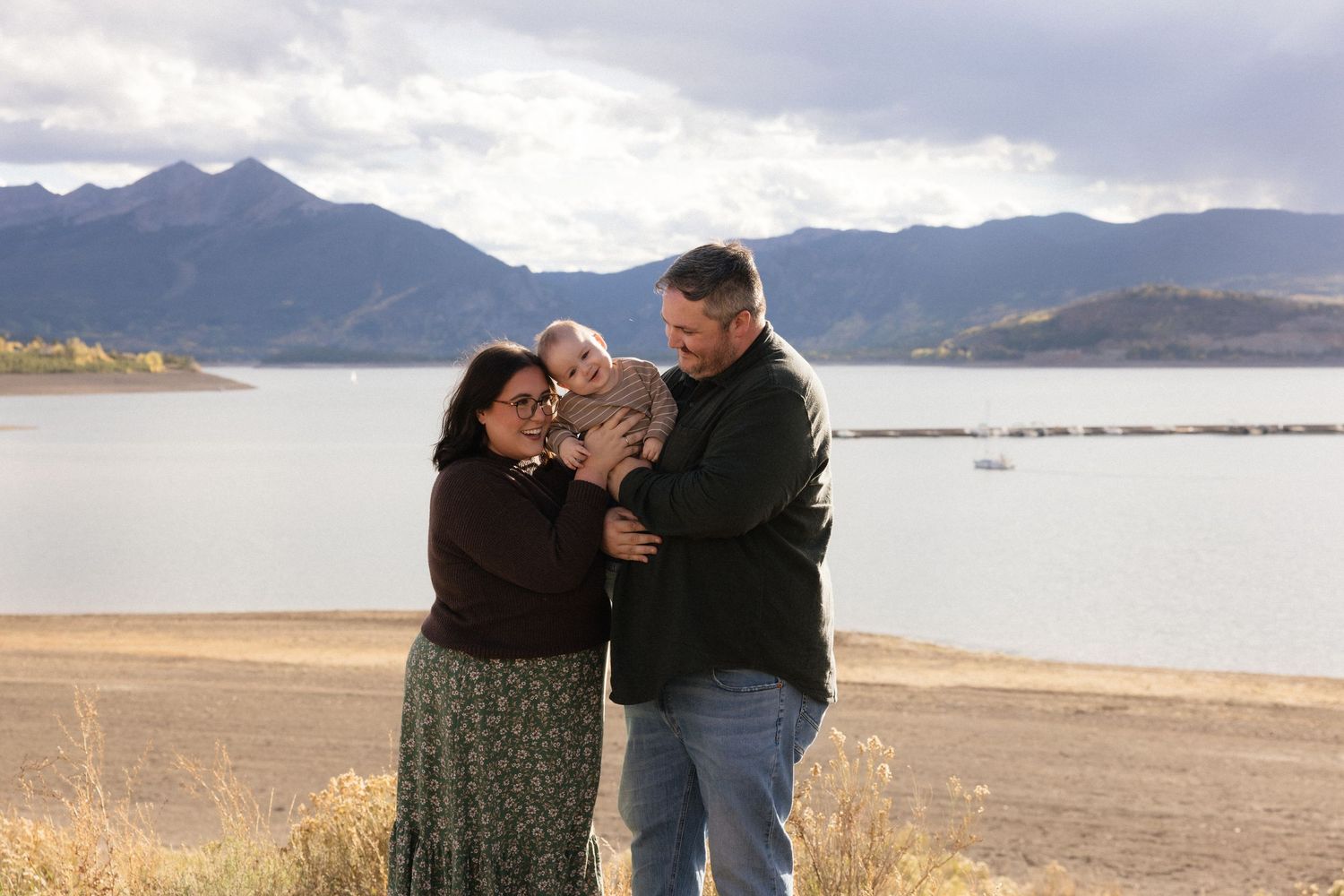 Family of three with baby posing near a mountain lake under cloudy skies in a scenic outdoor setting.