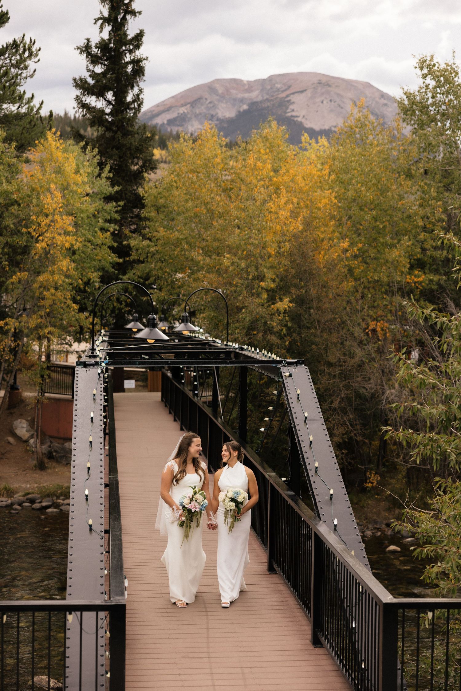 Two brides in white gowns walk hand-in-hand on a decorated bridge surrounded by autumn trees and mountains.