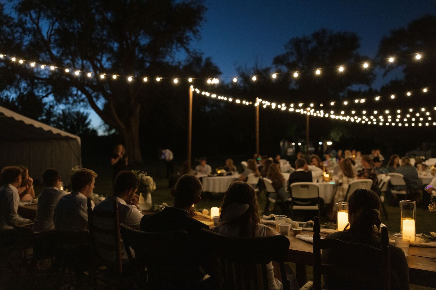Outdoor evening reception with string lights illuminating guests dining at tables under trees at night.