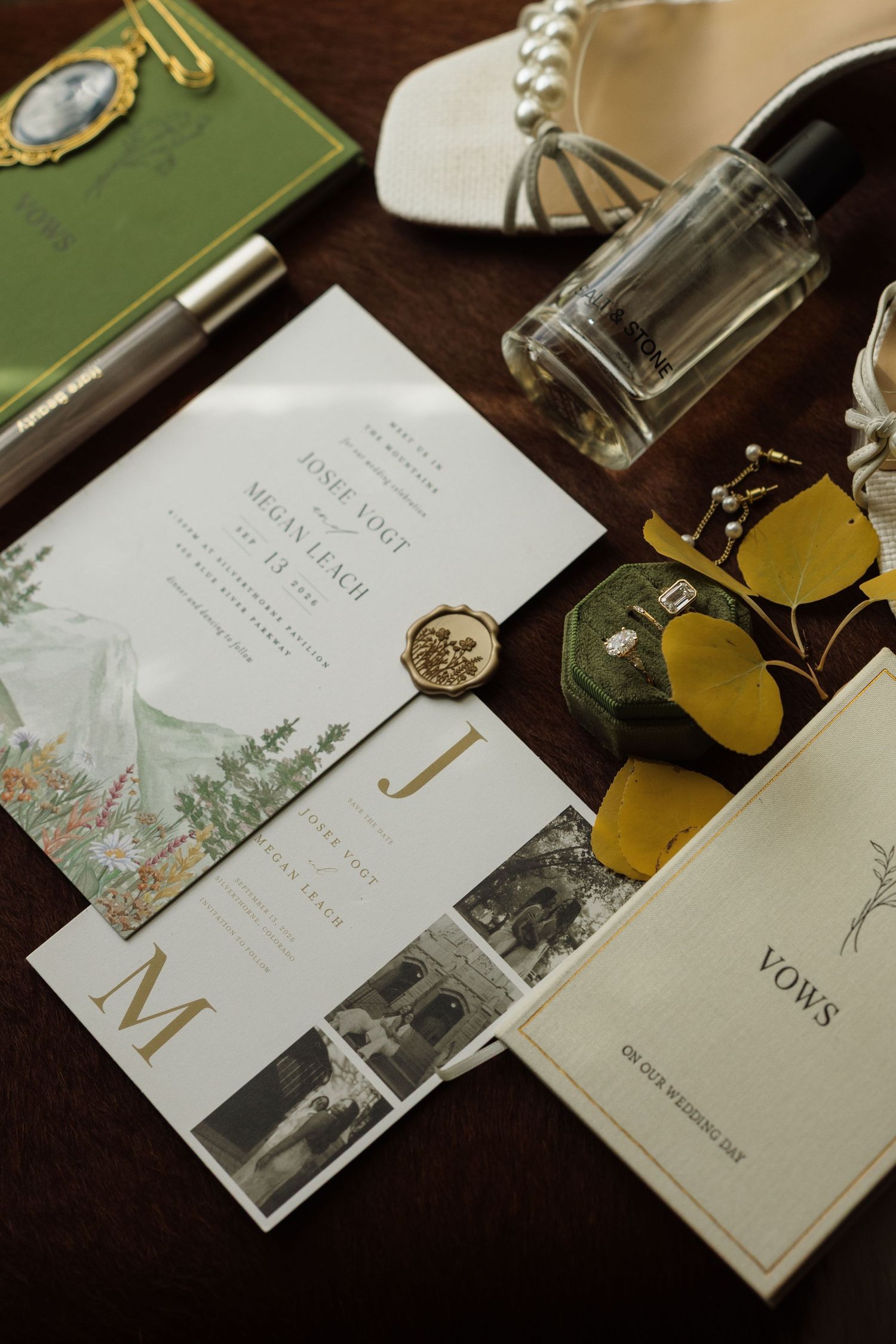 Elegant wedding flat lay with floral invitations, gold wax seal, vows booklet, perfume, pearl heels, and green foliage on dark wood.