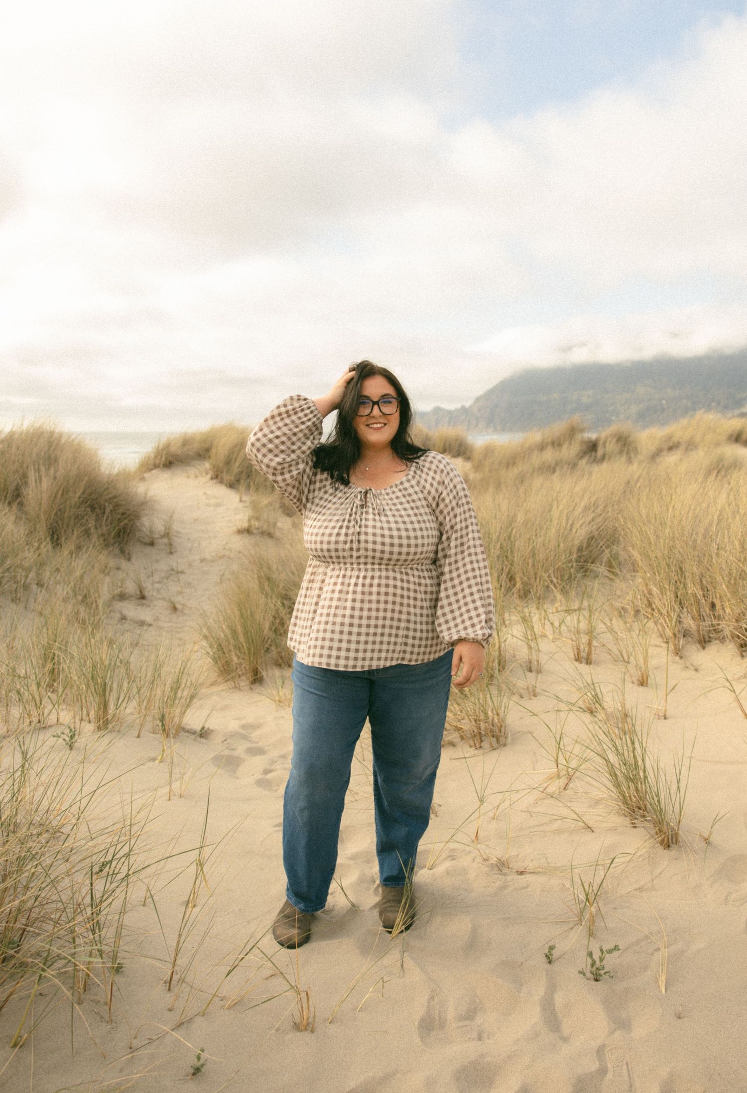 Person wearing gingham top and jeans poses playfully in sandy beach dunes at golden hour.