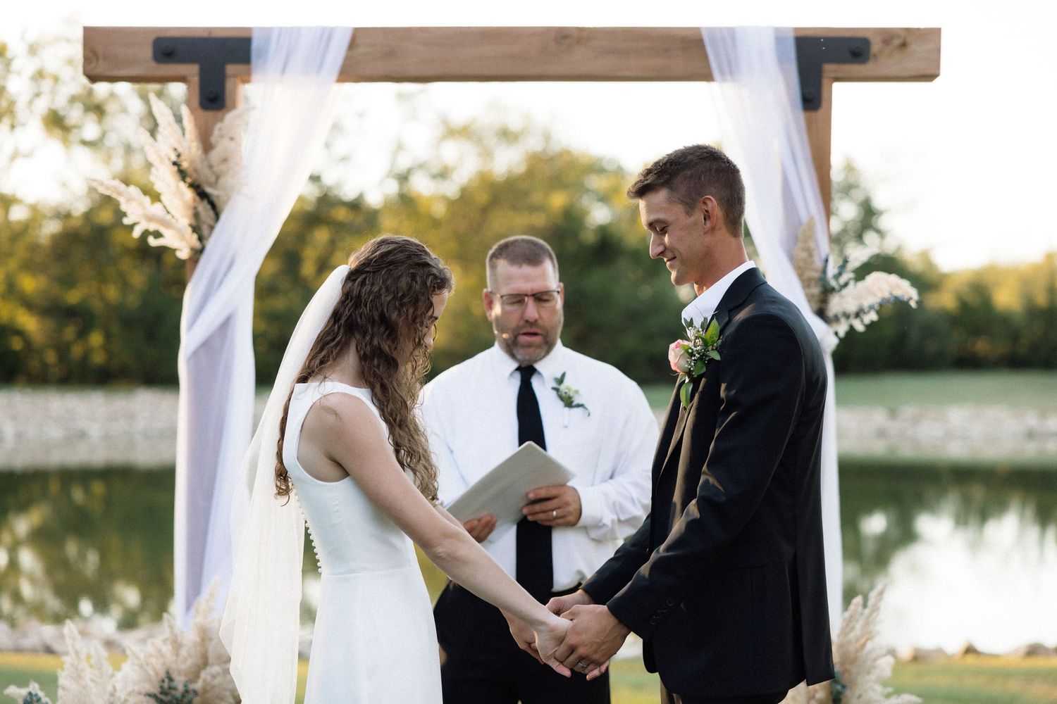 Couple exchanges vows at outdoor wedding ceremony under wooden arch with white draping near a pond.