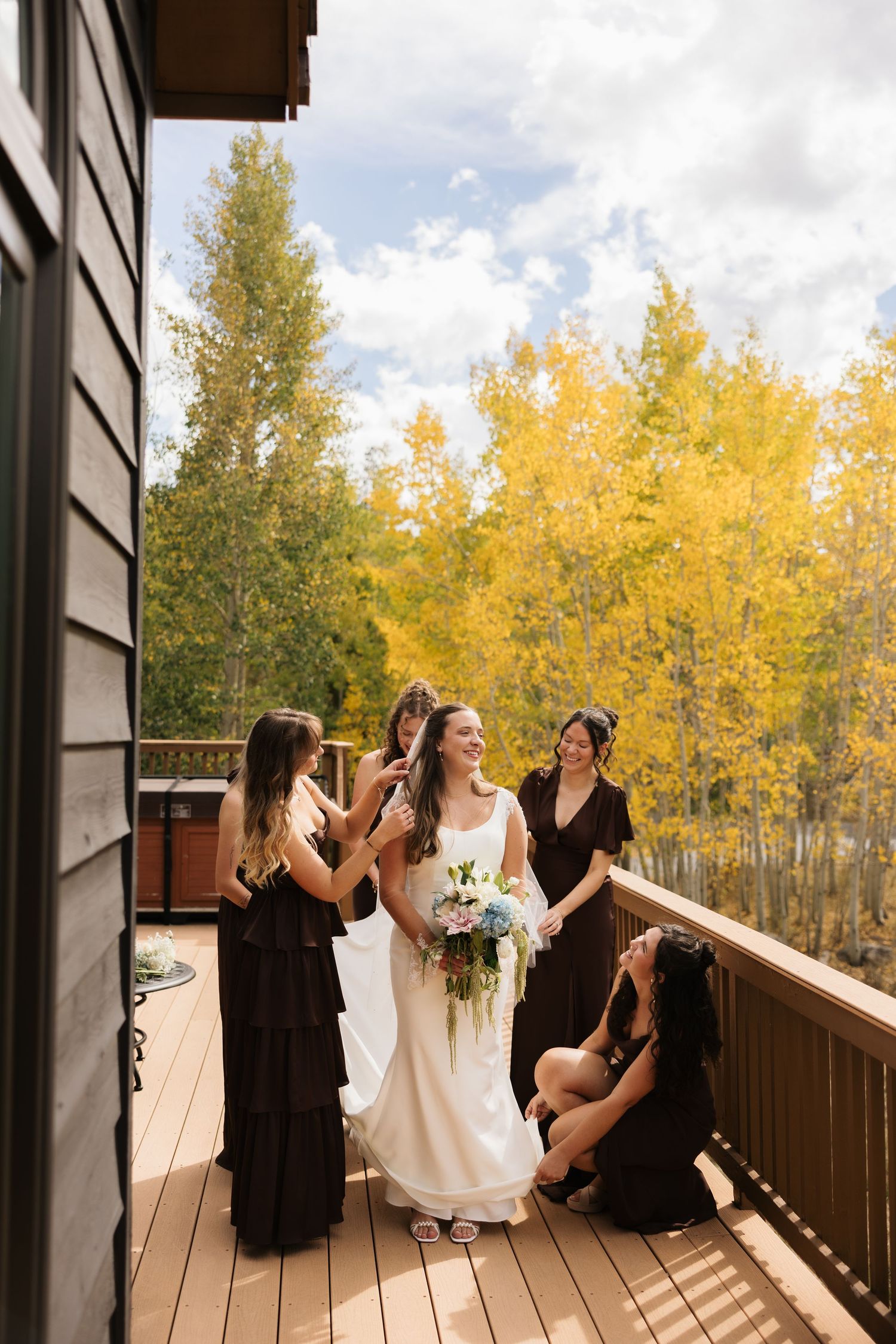 Bride in white gown holds bouquet on wooden deck with bridesmaids in dark dresses, surrounded by golden autumn trees.