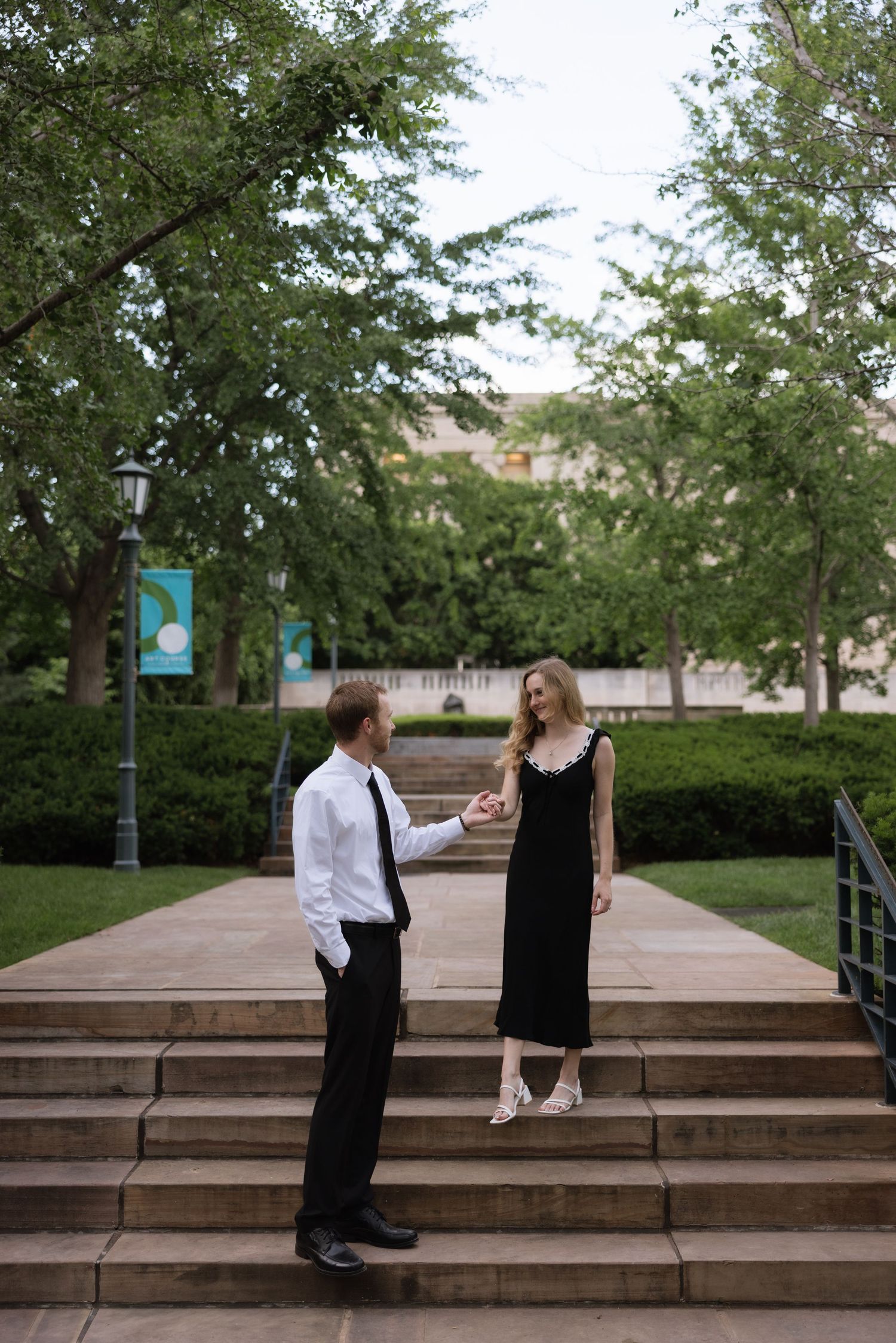 A couple in formal attire holds hands on outdoor steps surrounded by lush green trees and manicured hedges.