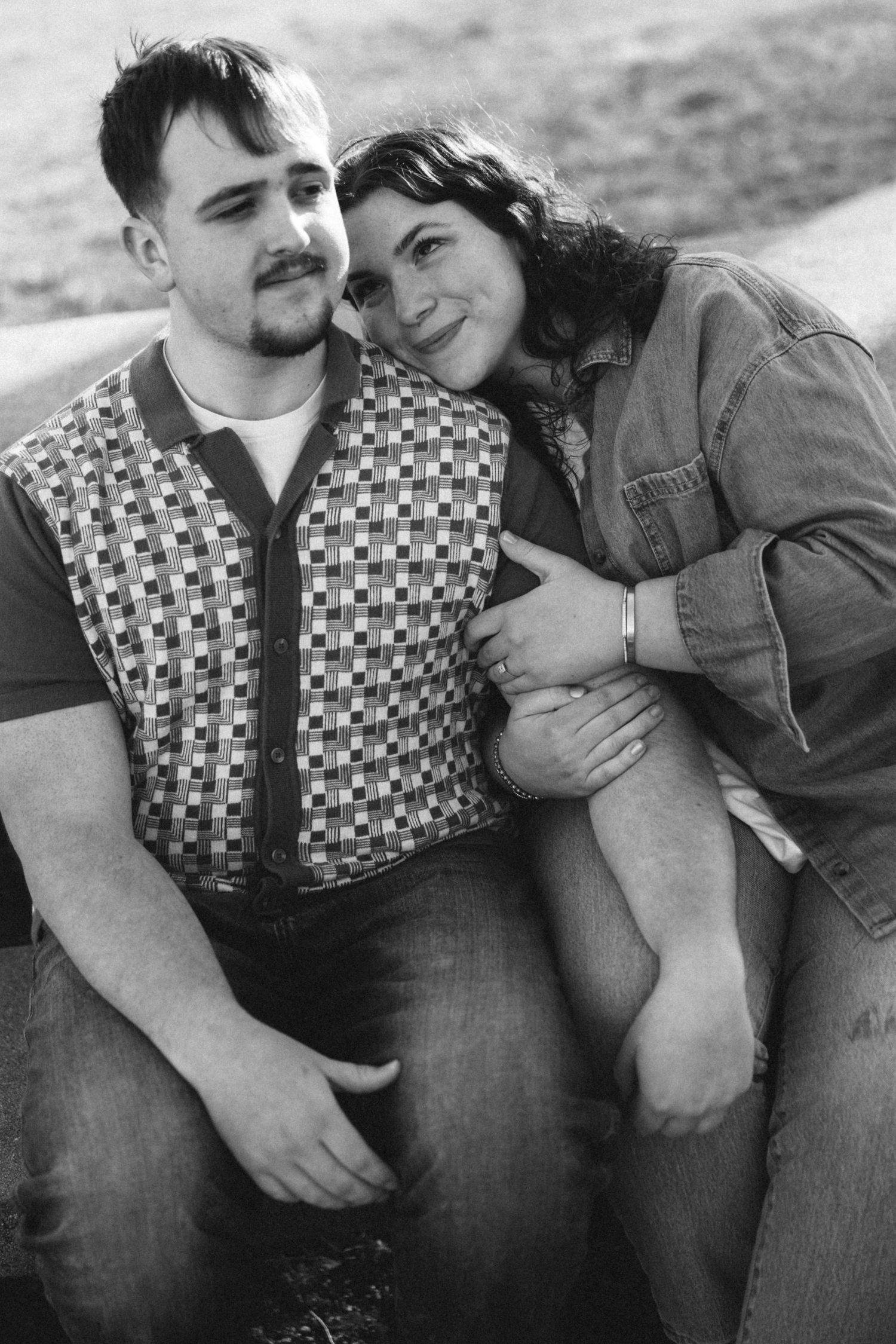 Black and white photo of a smiling couple sitting closely together outdoors, woman leaning on mans shoulder affectionately.