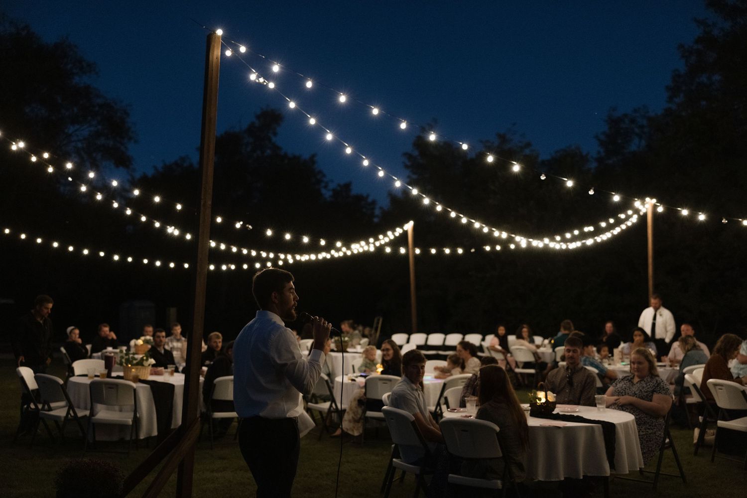 Outdoor evening wedding reception with string lights draped over tables where guests dine under a twilight sky.