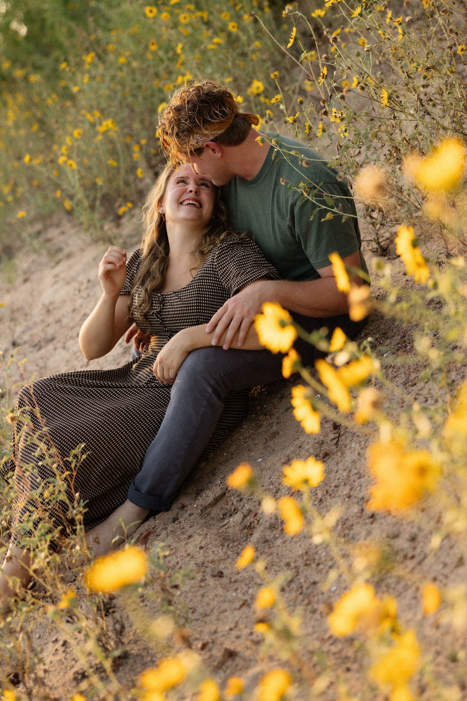 Couple sitting among yellow wildflowers, man kissing woman's head as she laughs joyfully outdoors.