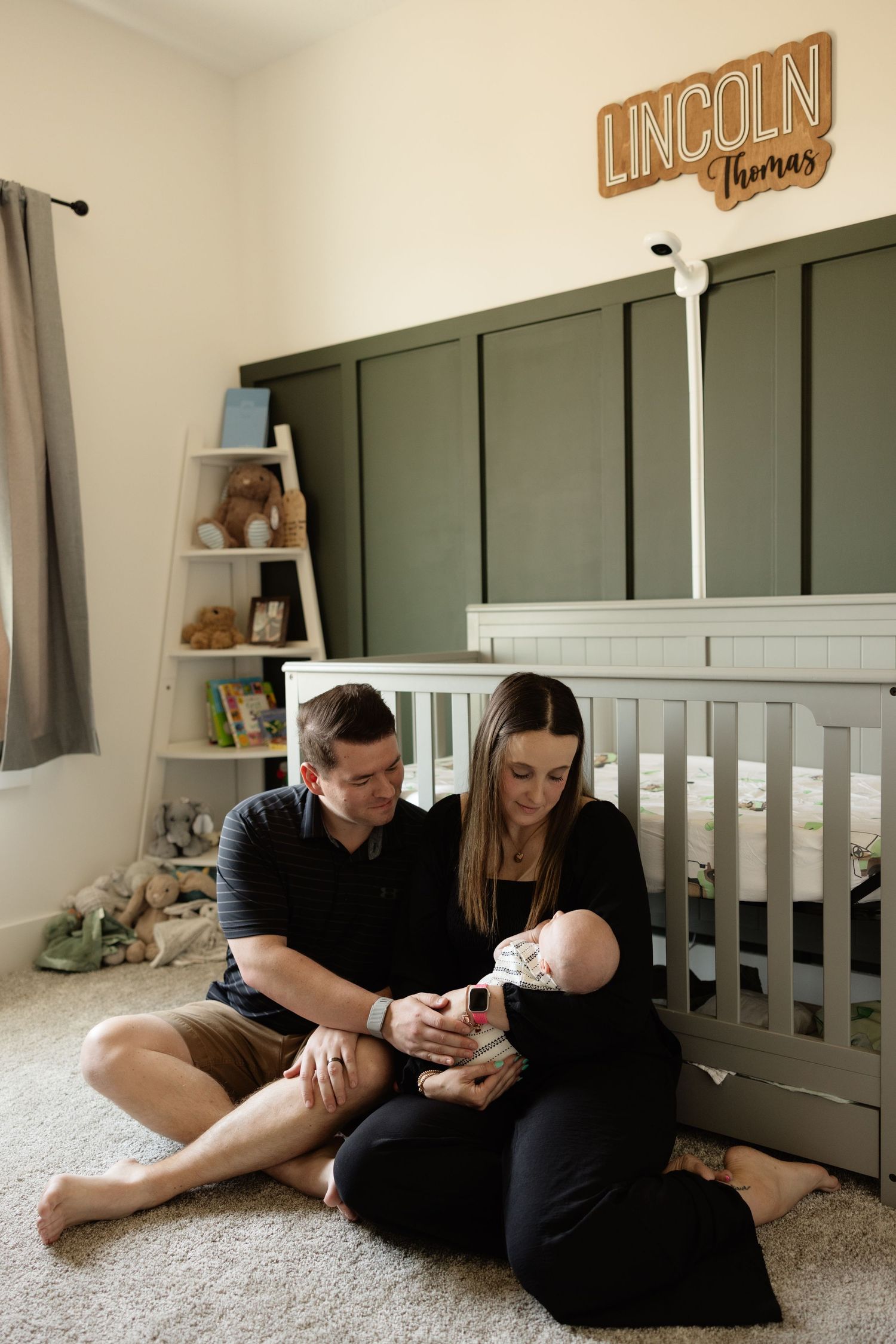 New parents sit on nursery floor holding newborn baby in front of gray crib below Lincoln Thomas name sign.