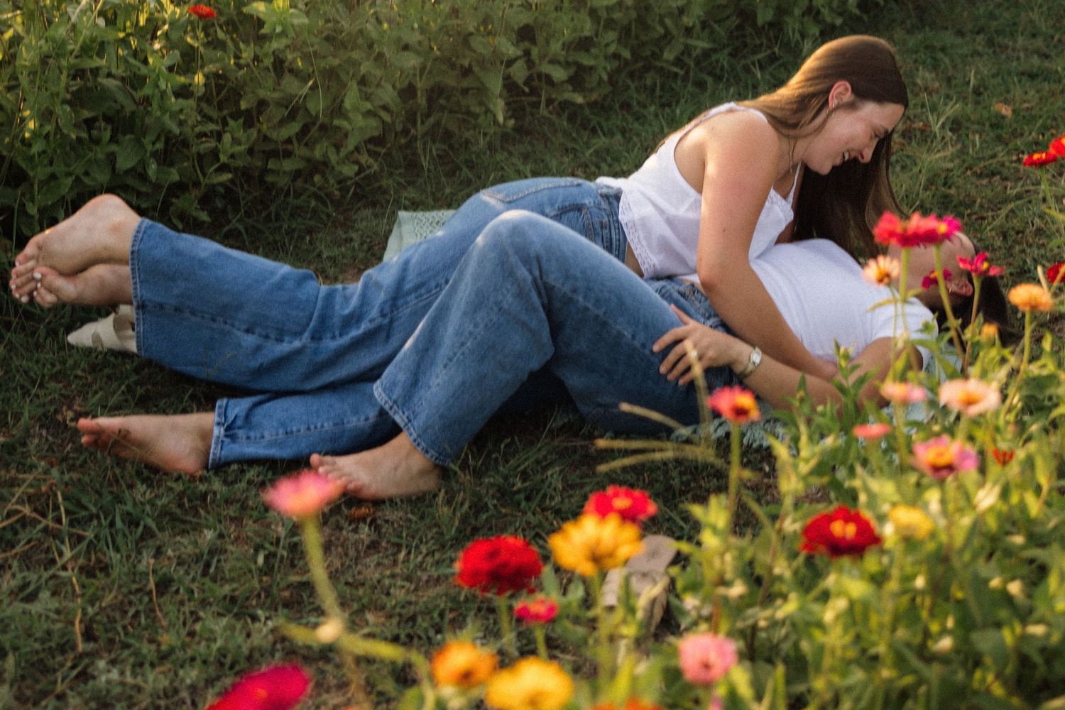 Two women relaxing together in a flower garden, lying on green grass surrounded by colorful blooms.