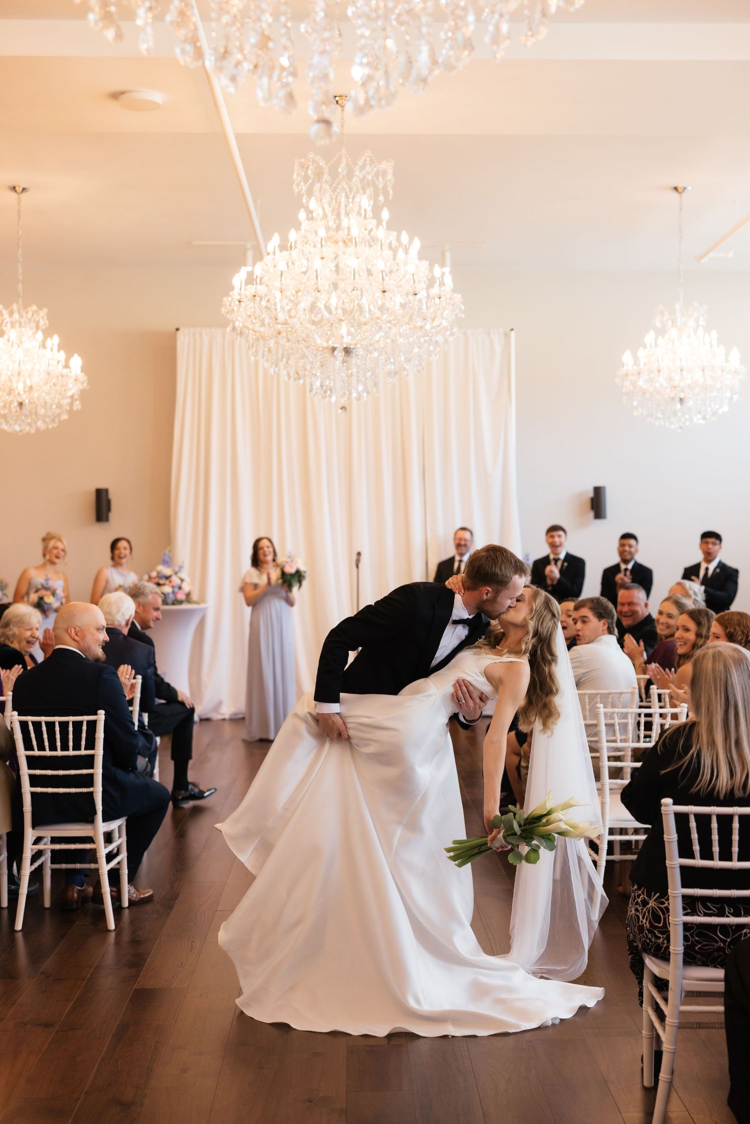 Groom dips bride for a kiss at elegant wedding ceremony with crystal chandeliers and white draped backdrop.