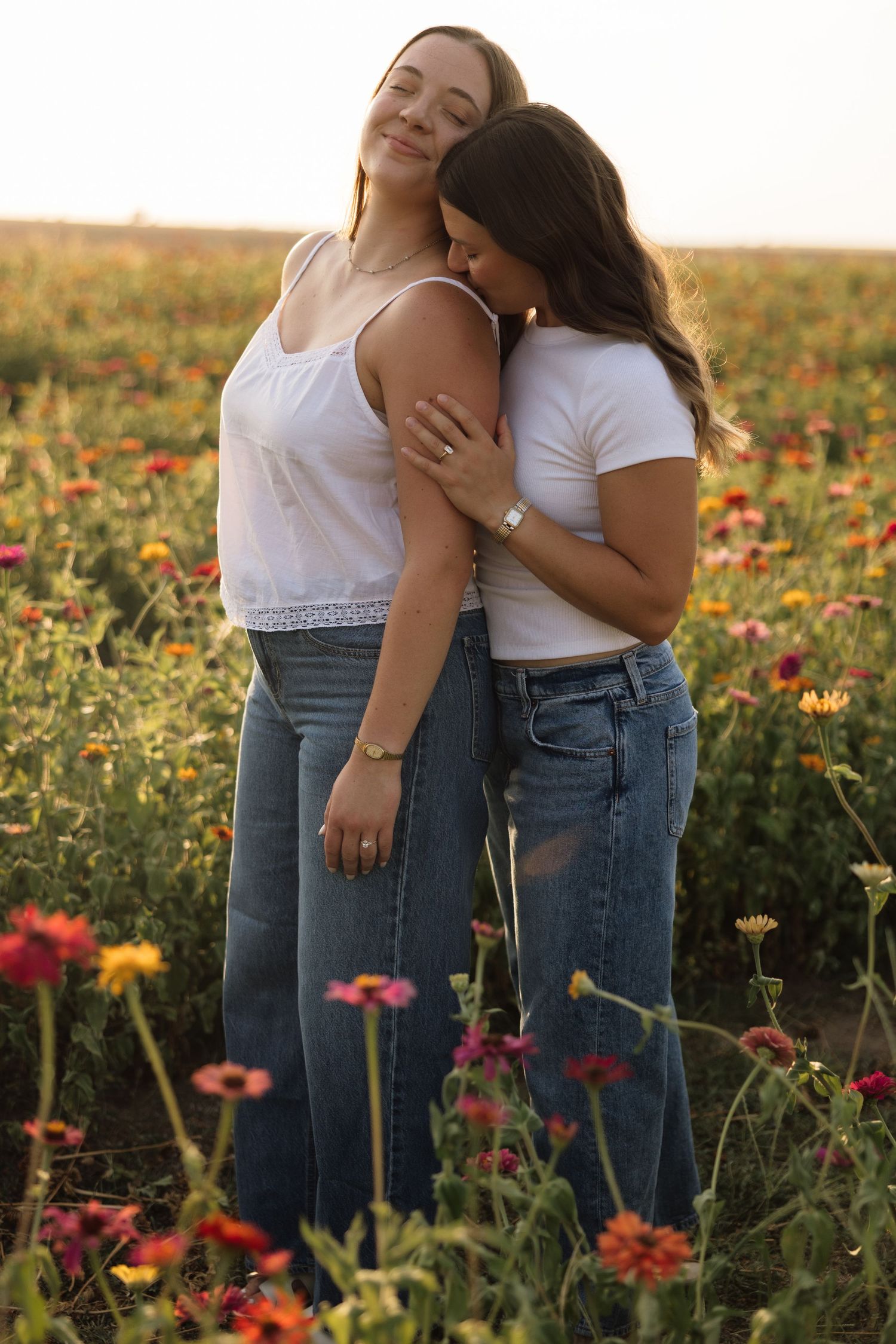 Two women in white tops and jeans embrace in a colorful flower field at golden hour.