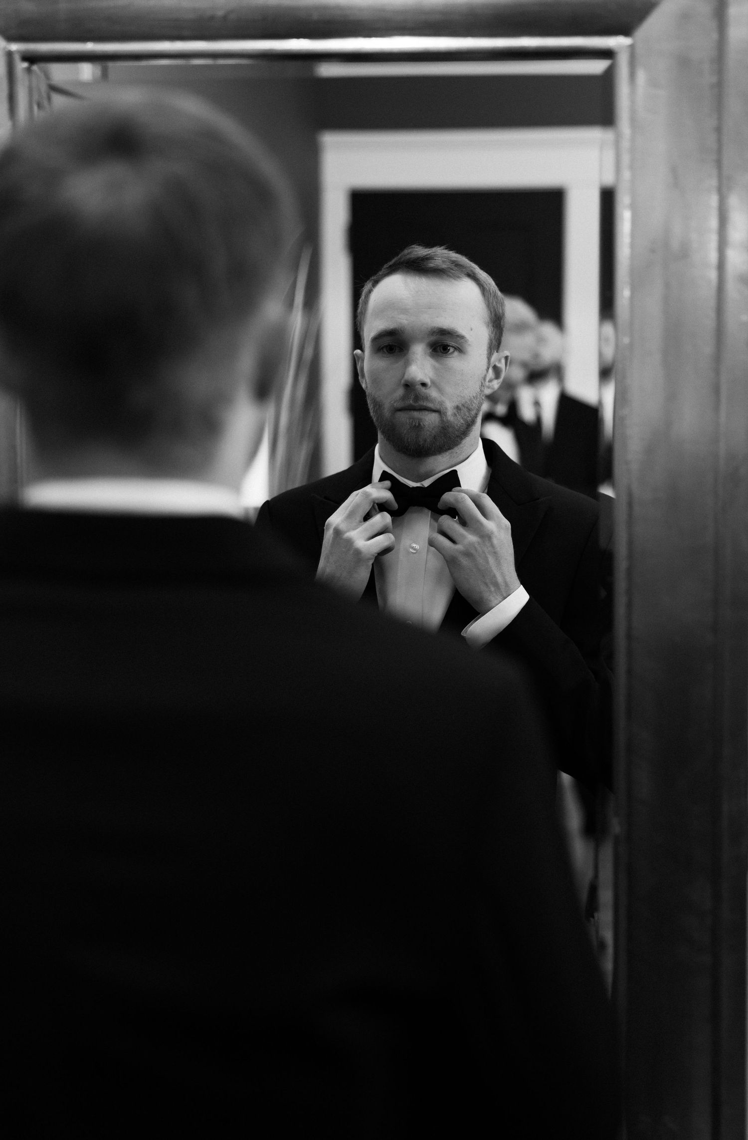 A man in a tuxedo adjusts his bow tie while looking at his reflection in a mirror, black and white photo.