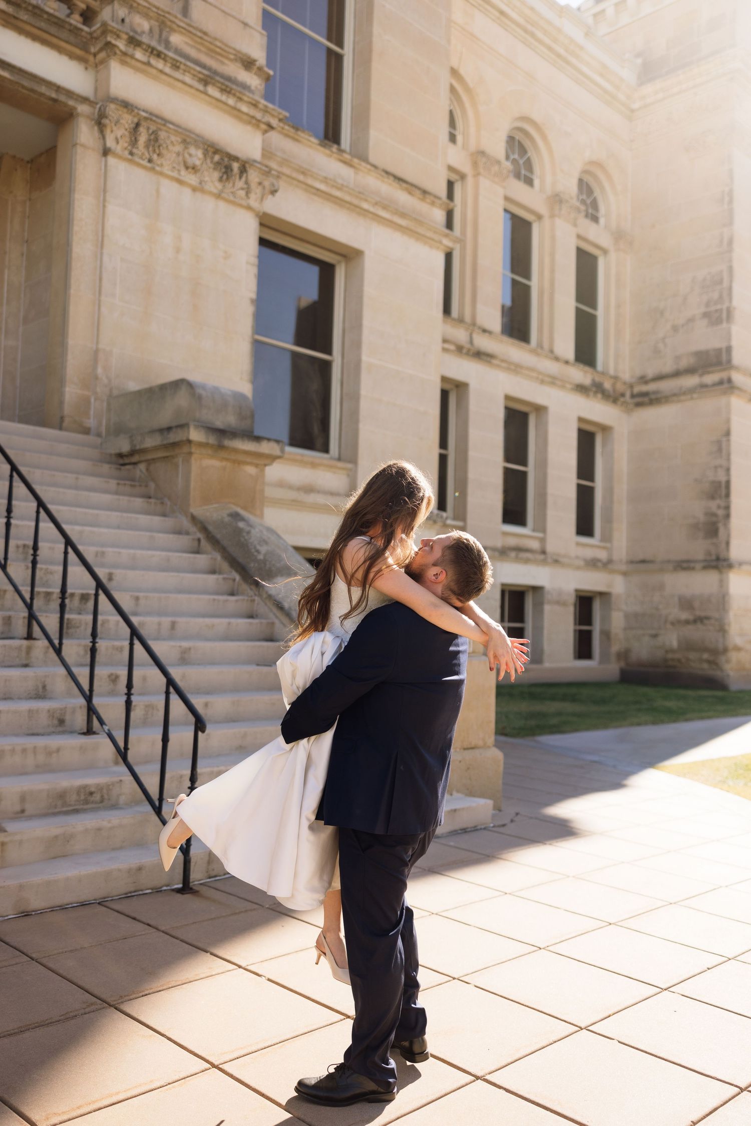 Romantic couple embraces and spins on stone steps outside a grand historic building in warm sunlight.