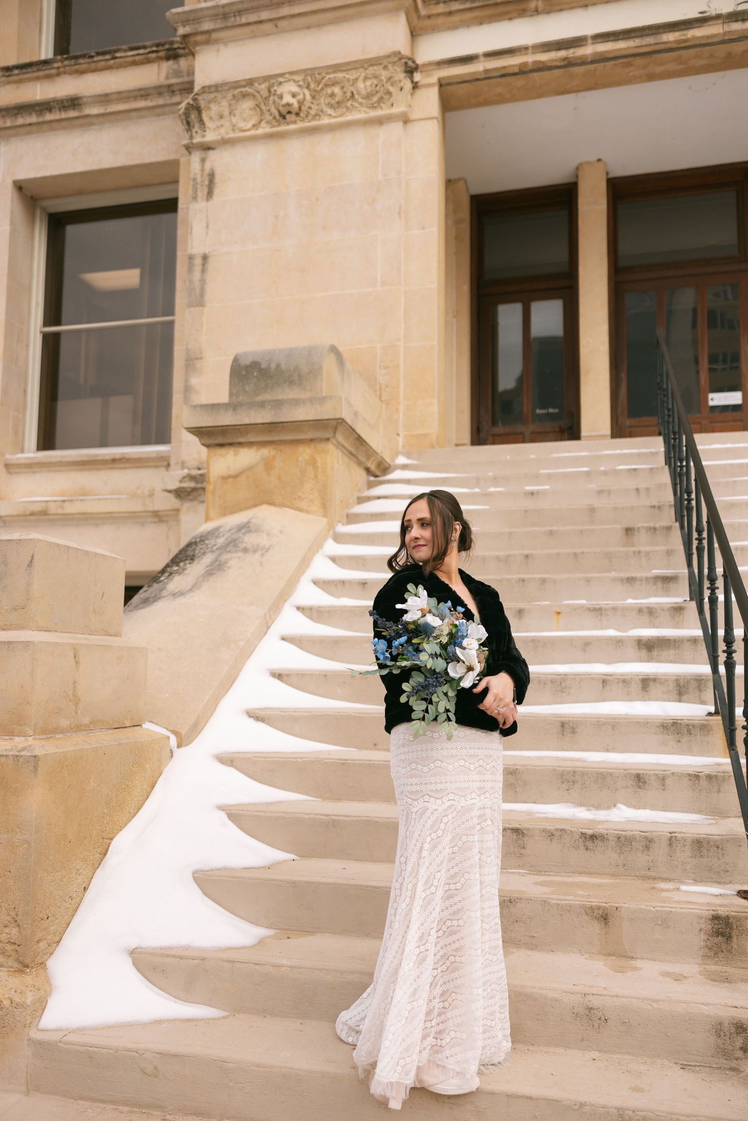 A bride in a white lace dress twirls on stone steps outside a historic building.