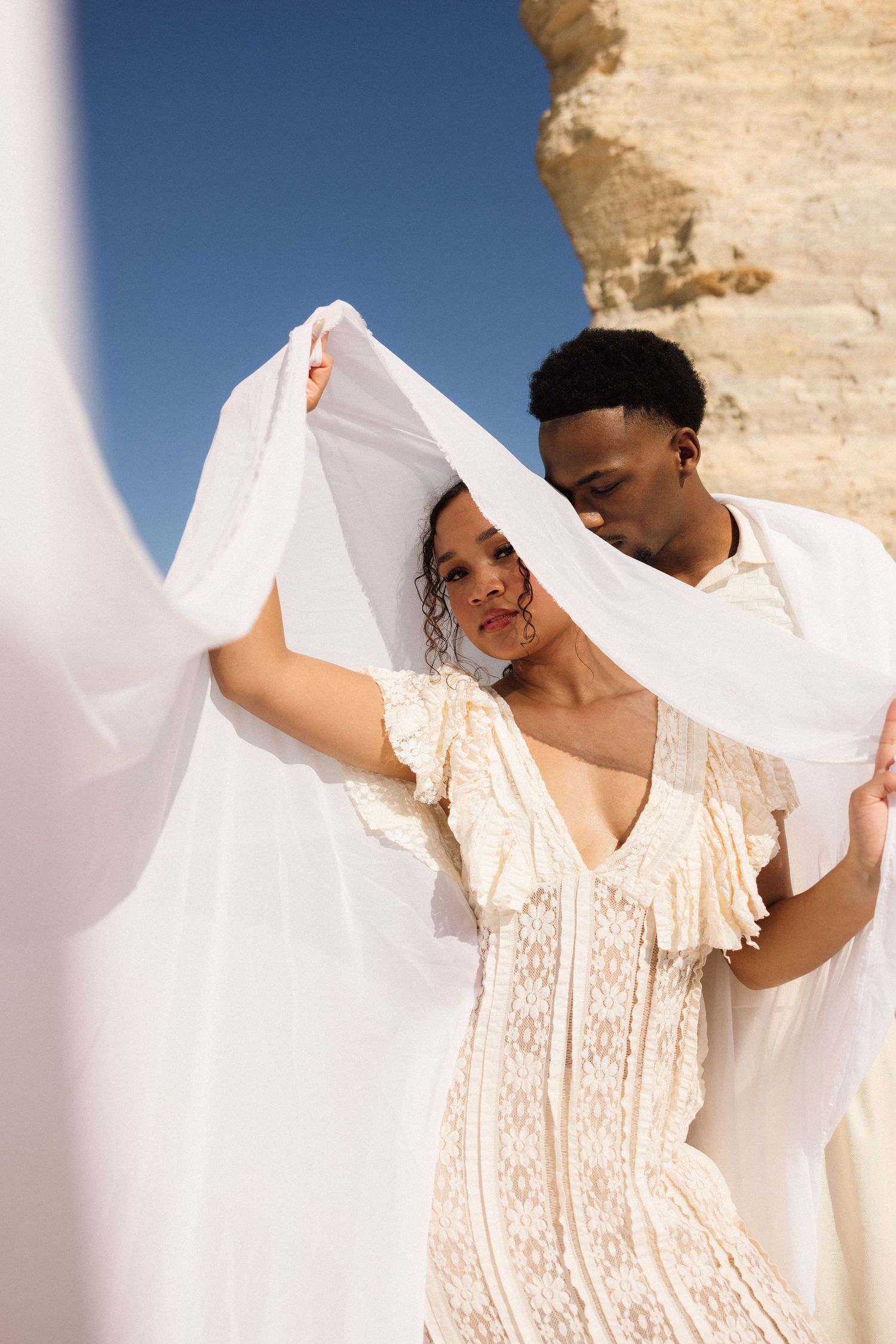 A couple in white attire poses under a flowing white fabric against a rocky backdrop and blue sky.