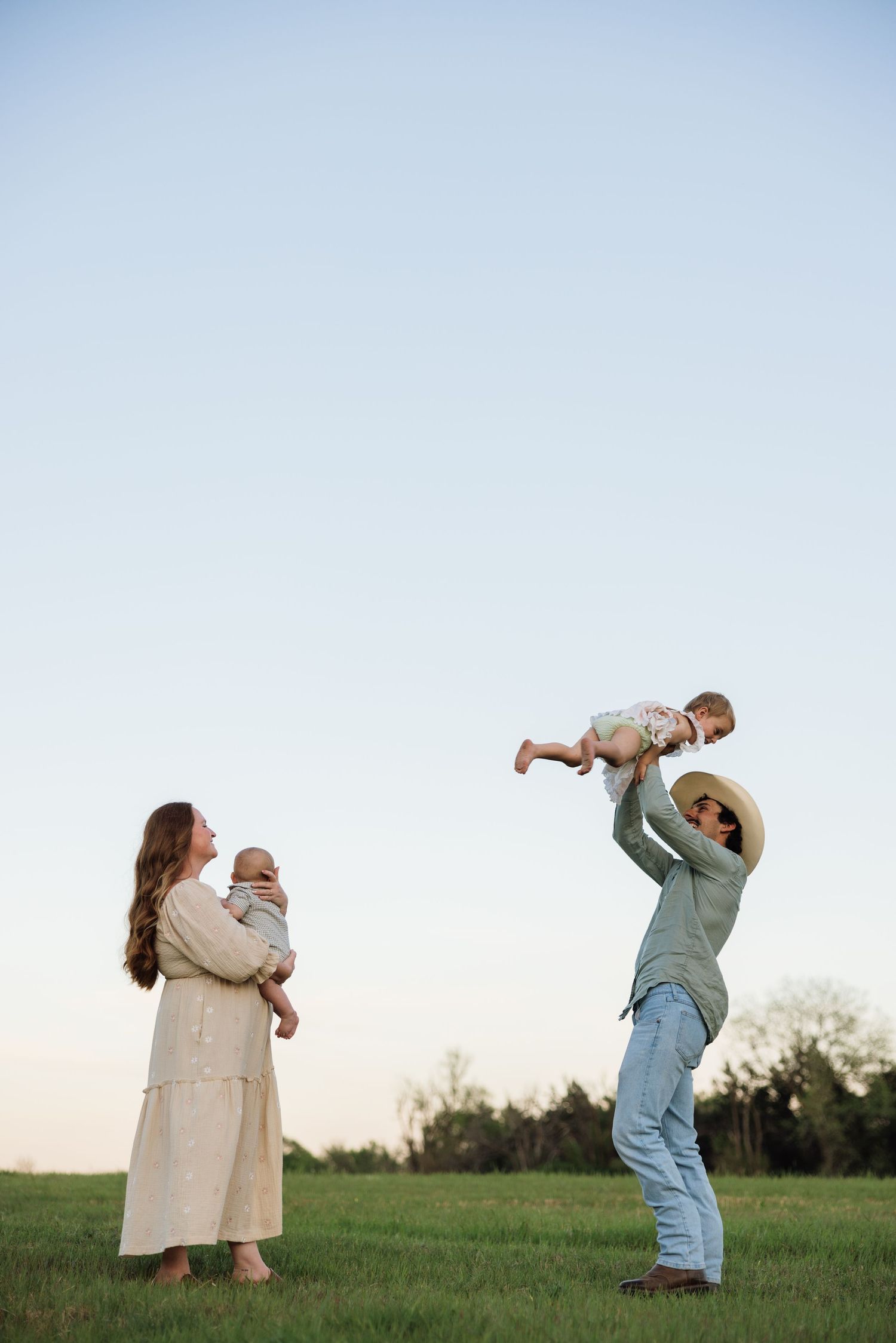 A family plays outdoors at dusk, mother holds baby while father lifts toddler high in a green field.