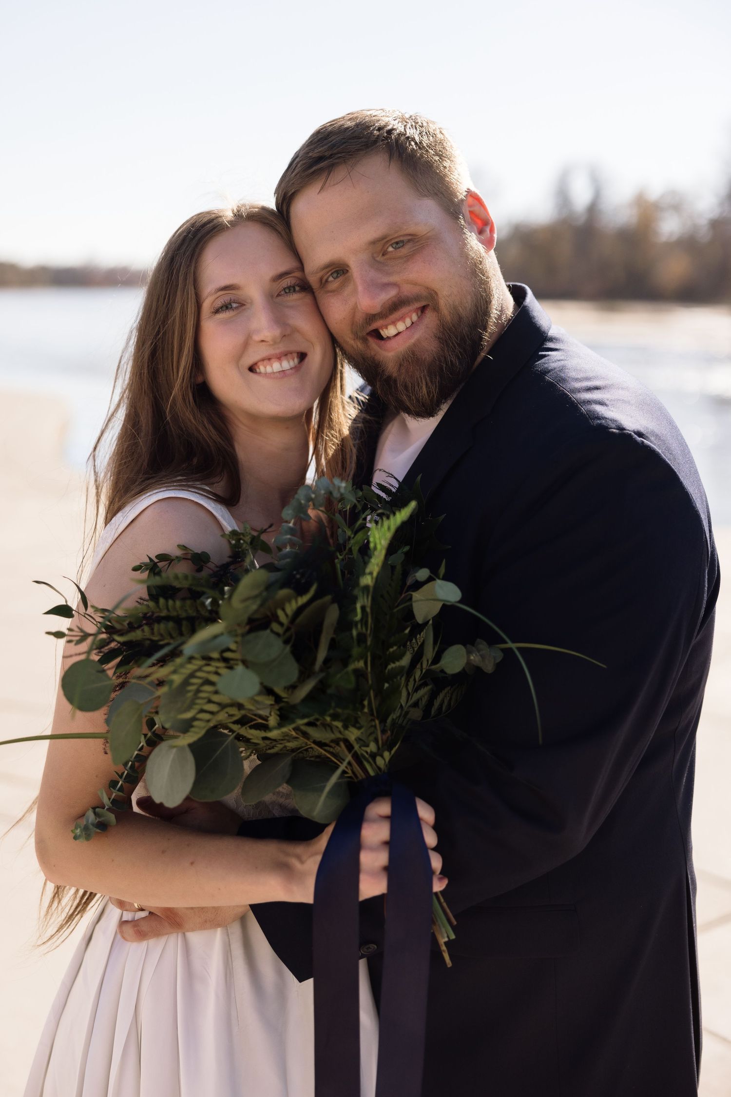 Winter beach wedding couple poses with greenery bouquet against bright background.