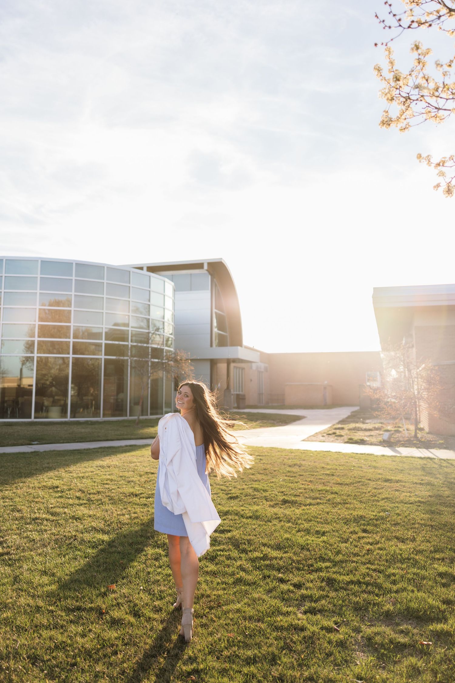 Person in white dress twirls in sunlit field near modern building.
