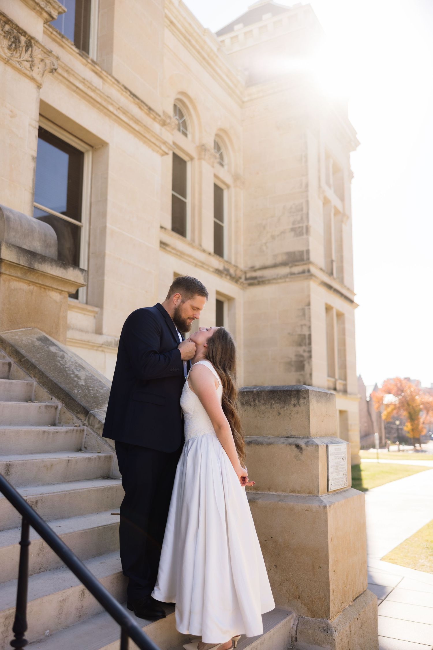 Wedding couple sharing a kiss on stone steps of historic building at sunset.
