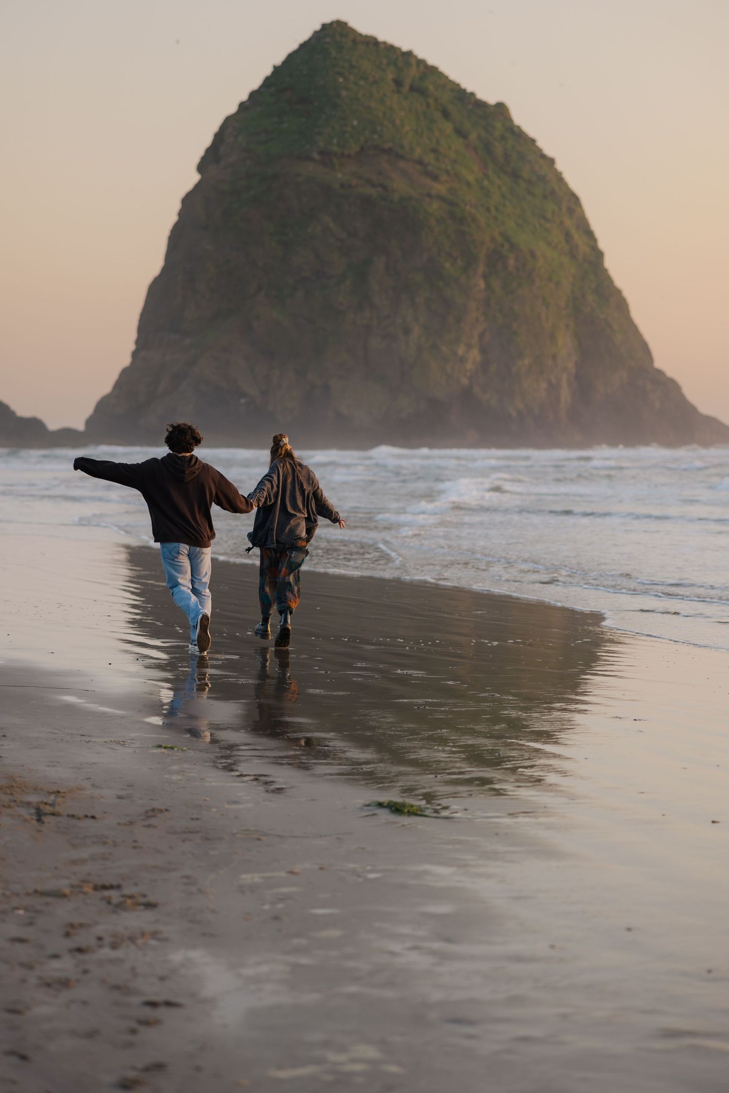 Two figures walking along a misty beach with large rock formation in background at sunset.