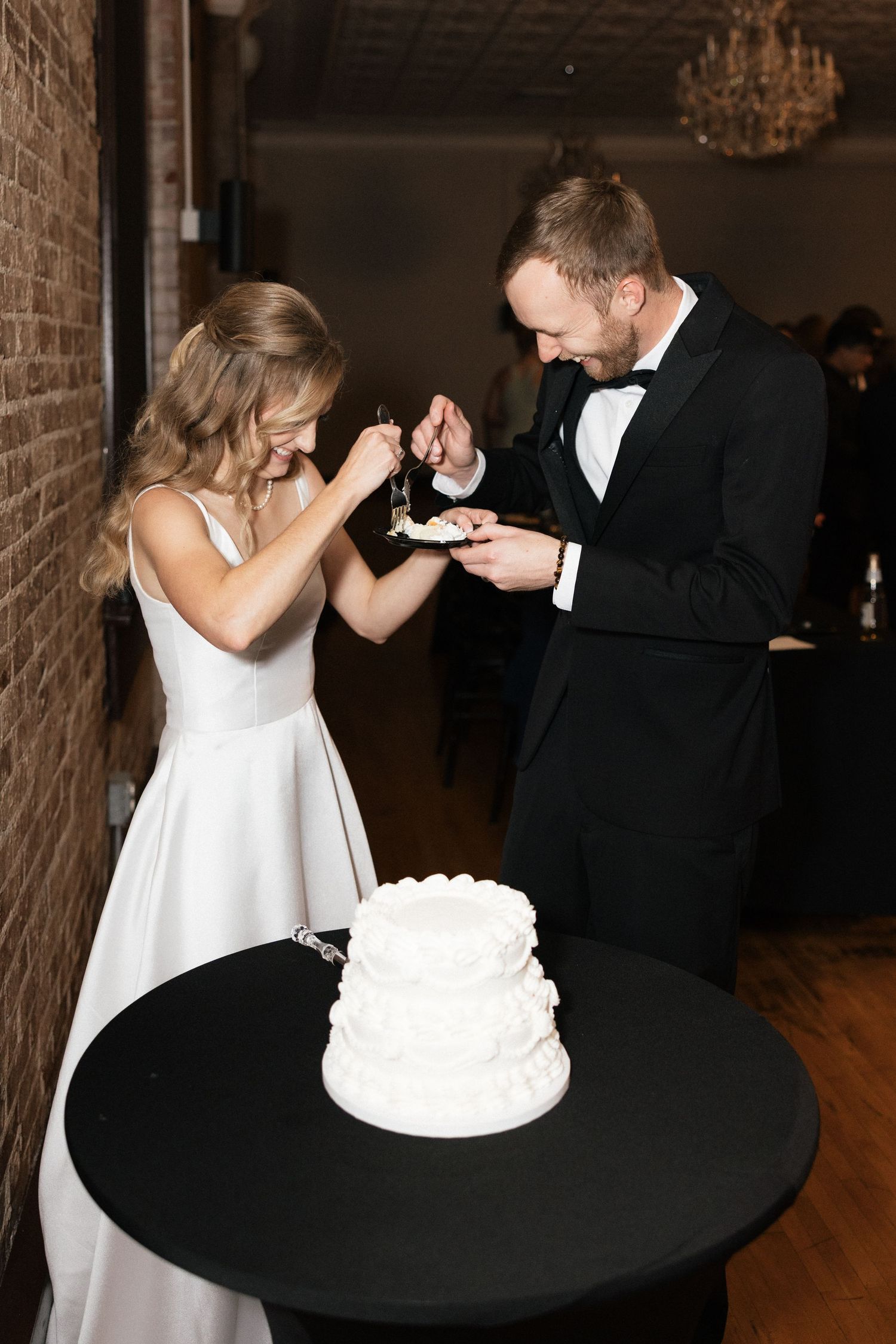 Bride and groom feeding each other wedding cake at elegant reception with white tiered cake on black table.