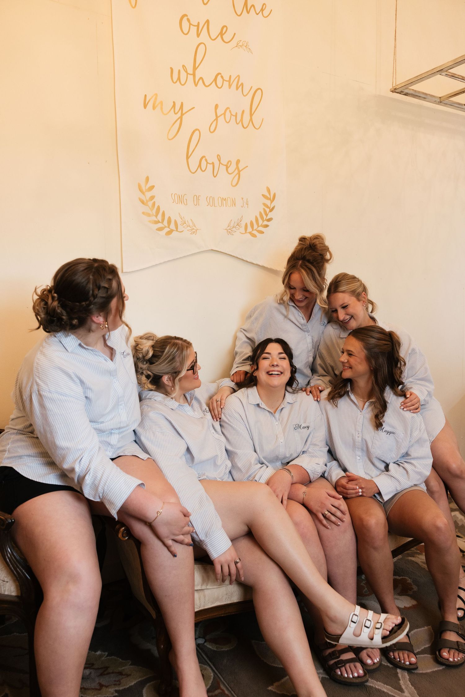 Bridesmaids laughing together in matching white button-up shirts seated on a couch with a romantic Song of Solomon wall sign.