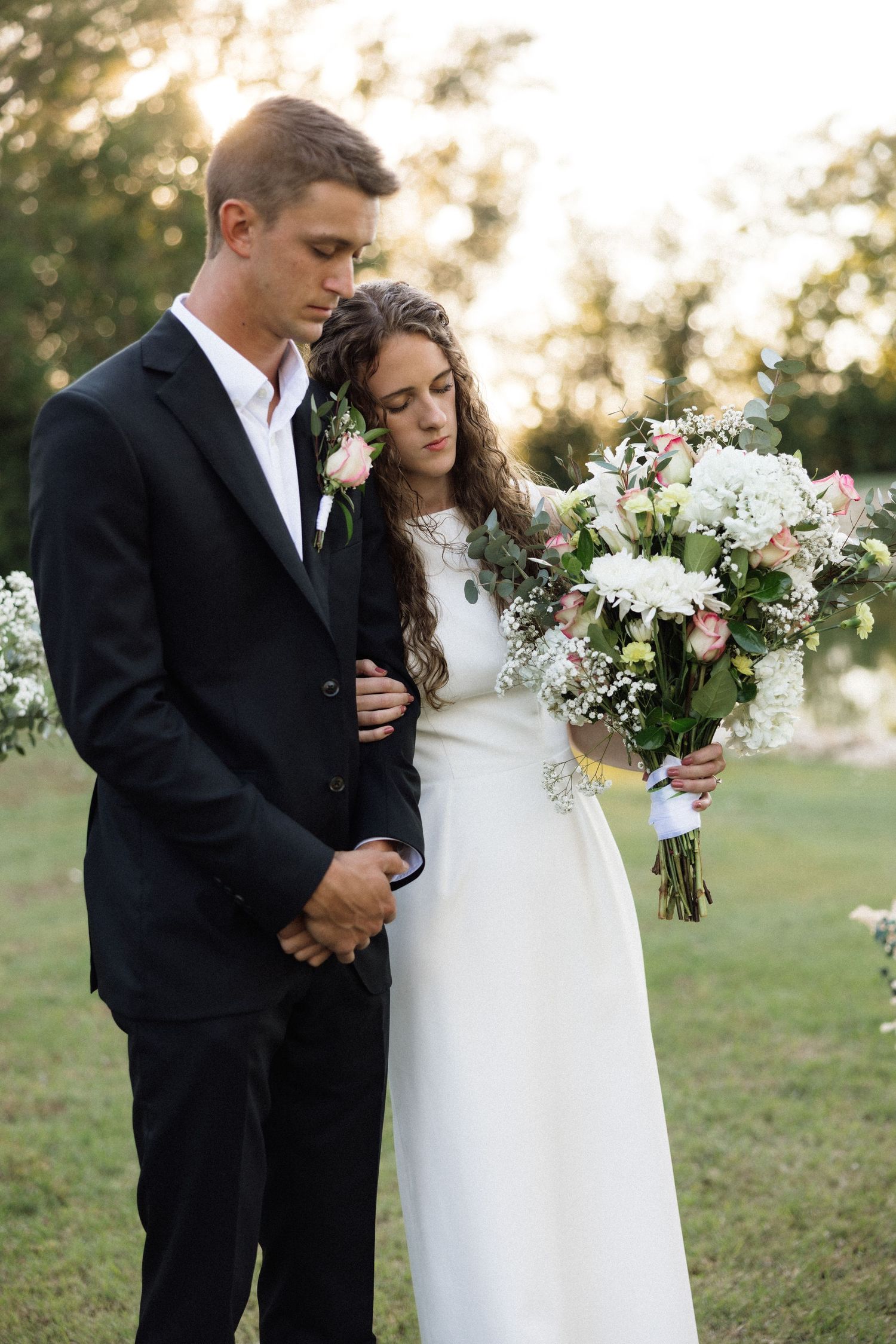 Bride in white gown holding floral bouquet stands beside groom in black suit during outdoor wedding ceremony.