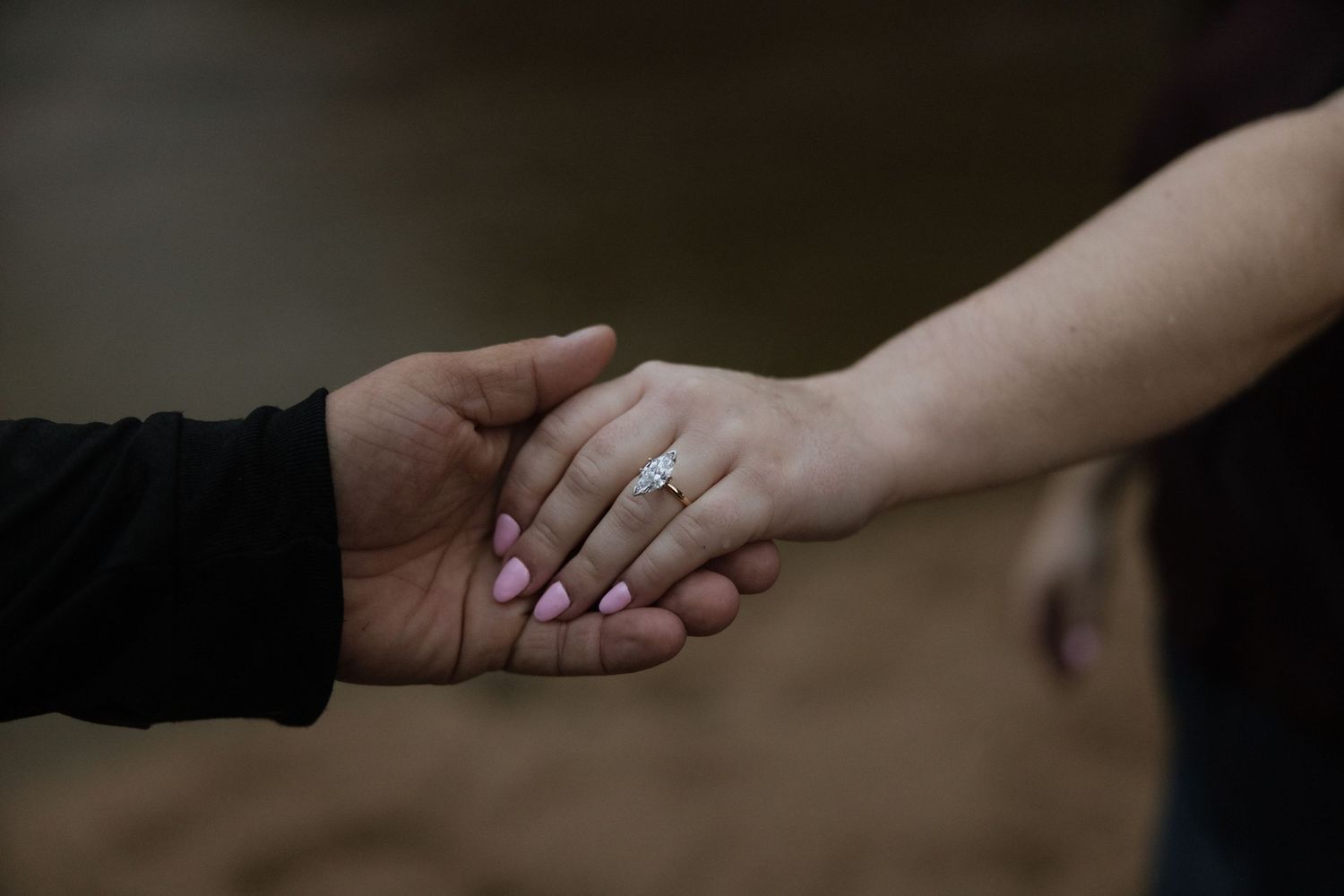 An interracial couple holds hands tenderly, showcasing a diamond engagement ring on the woman's finger with pink nails.