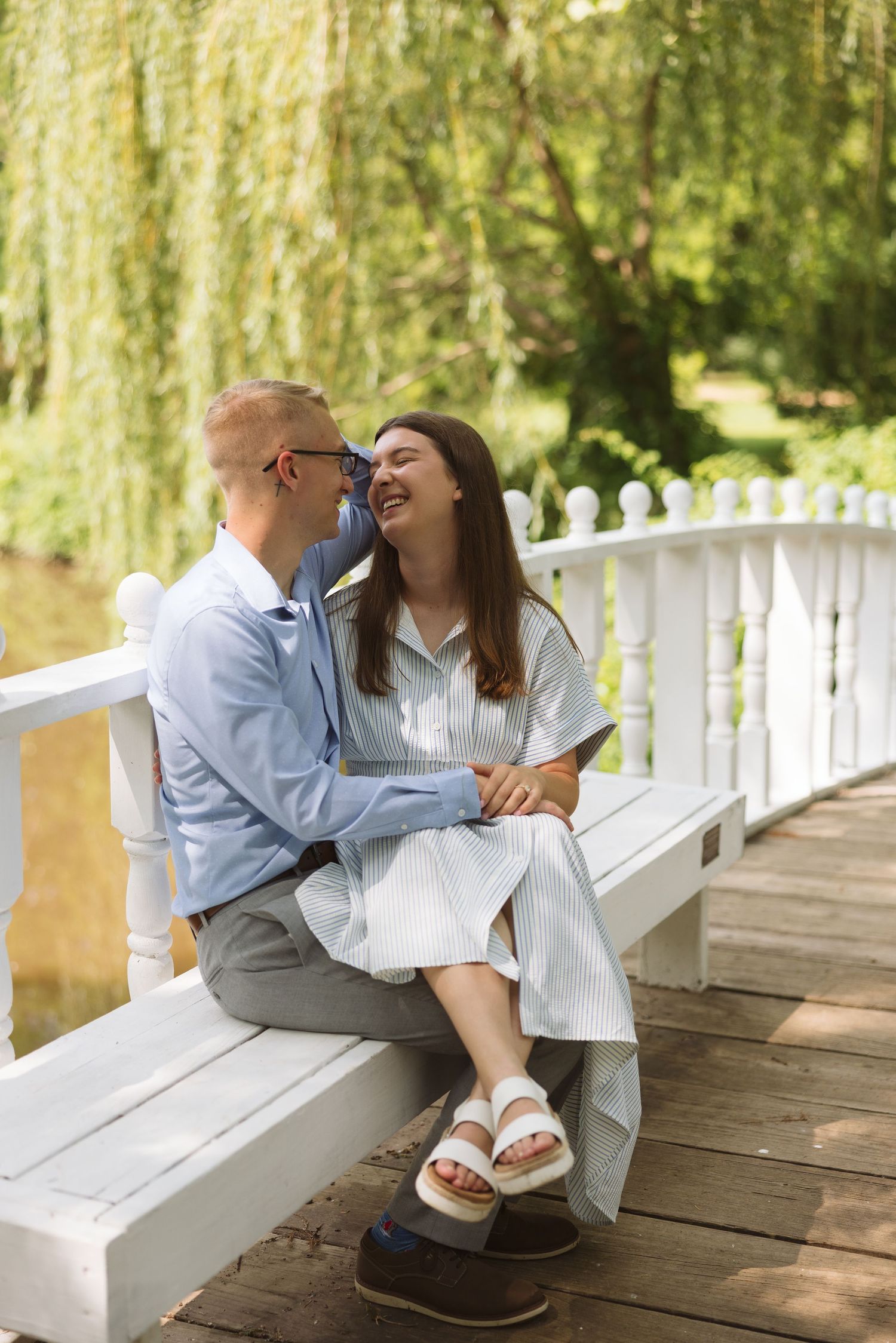 Couple shares intimate moment on white wooden bridge surrounded by willow trees.