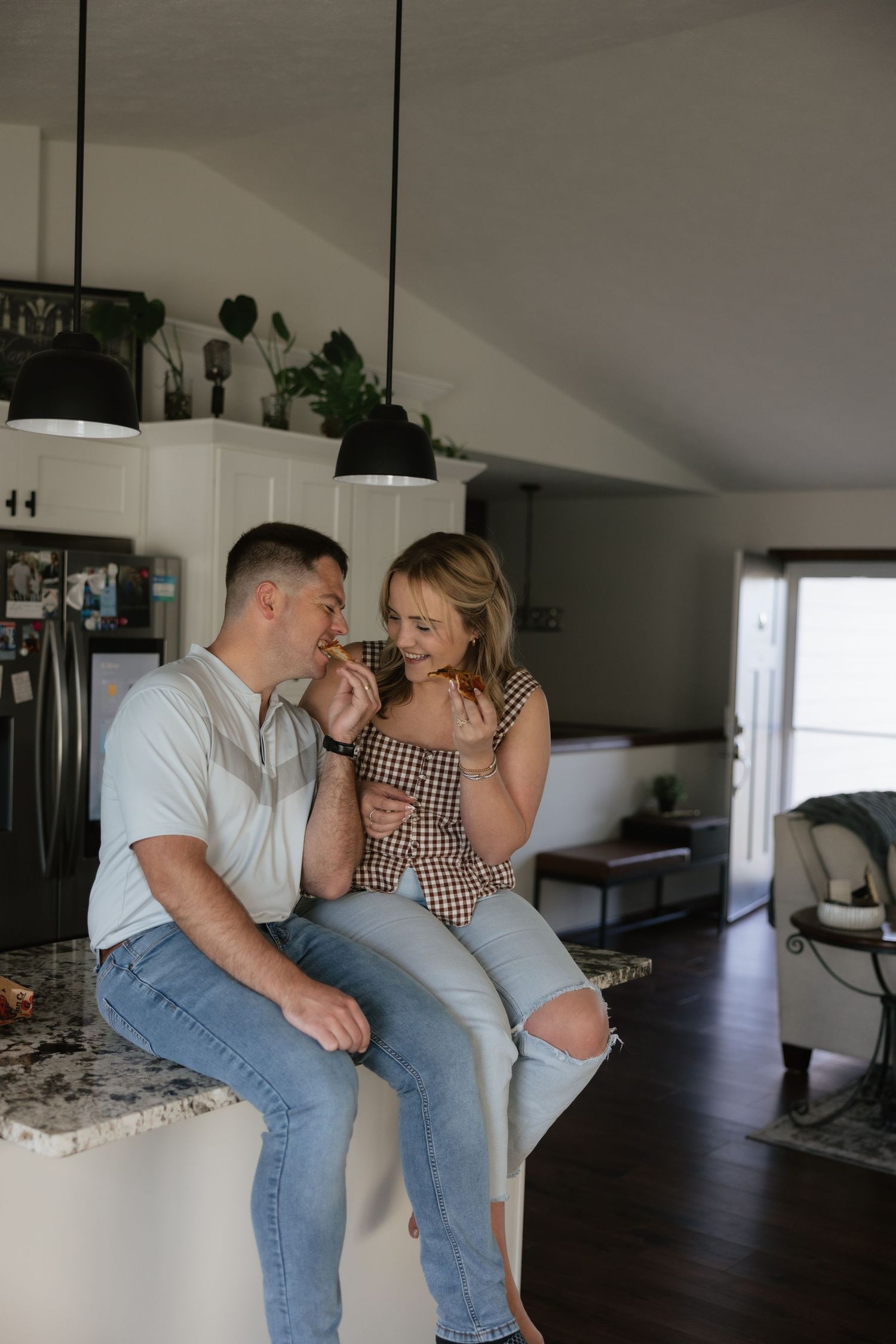 A couple shares a tender moment in a modern kitchen, sitting on a marble countertop beneath black pendant lights.