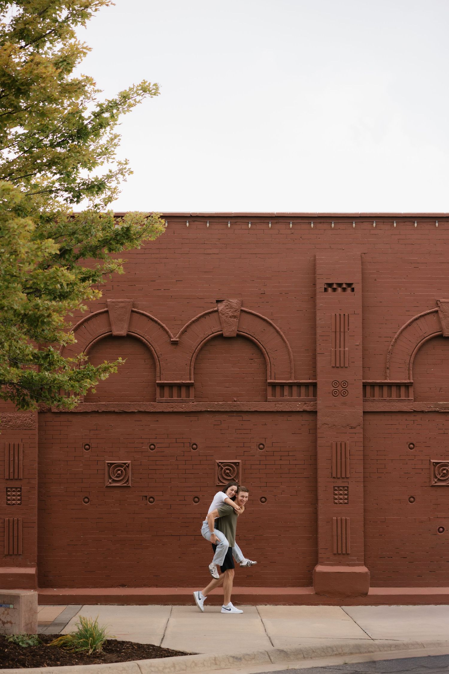 Couple sharing a piggyback ride on a sidewalk in front of a decorative red brick wall with arched details.