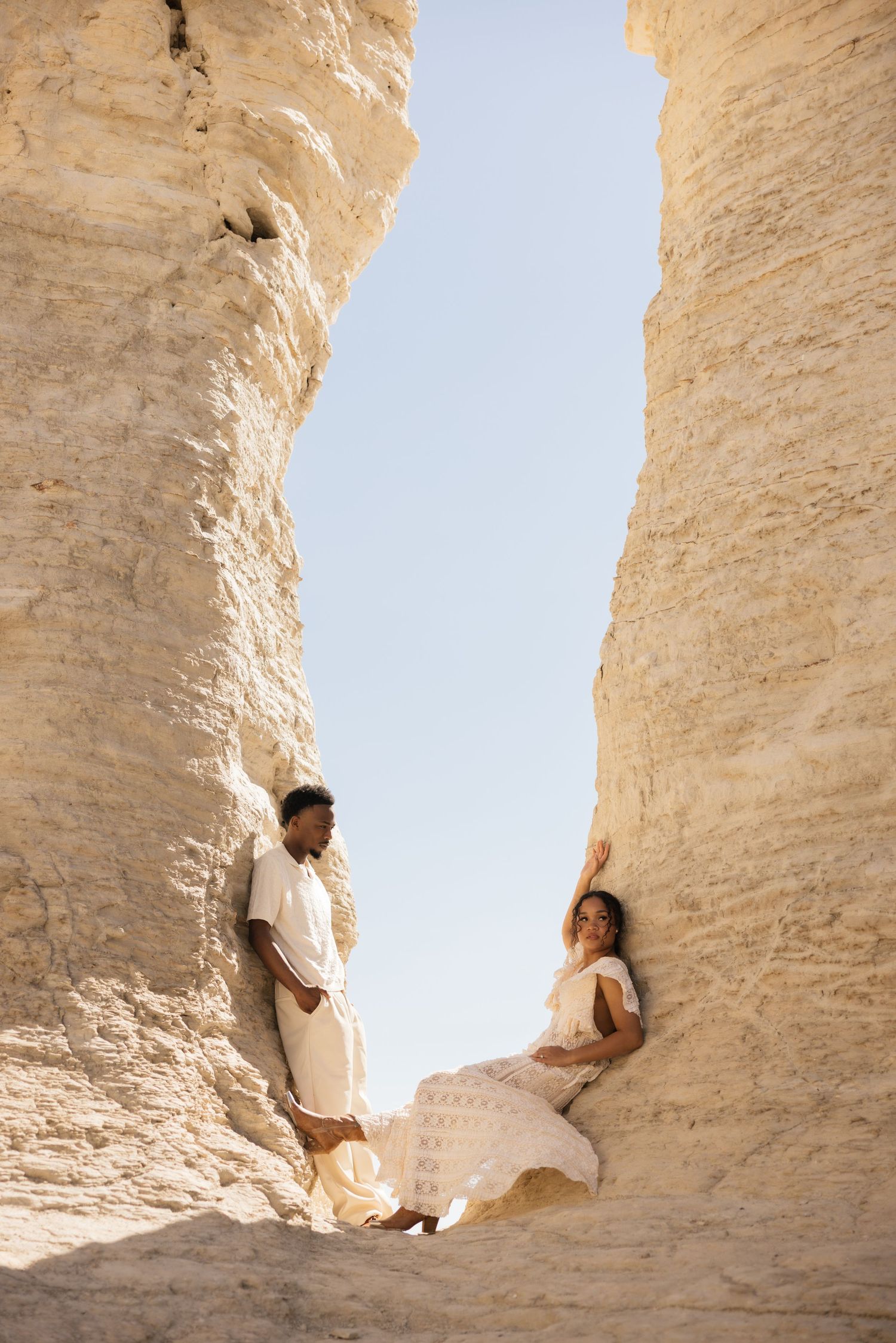 A couple in white outfits poses between towering sandstone rock formations under a clear blue sky.