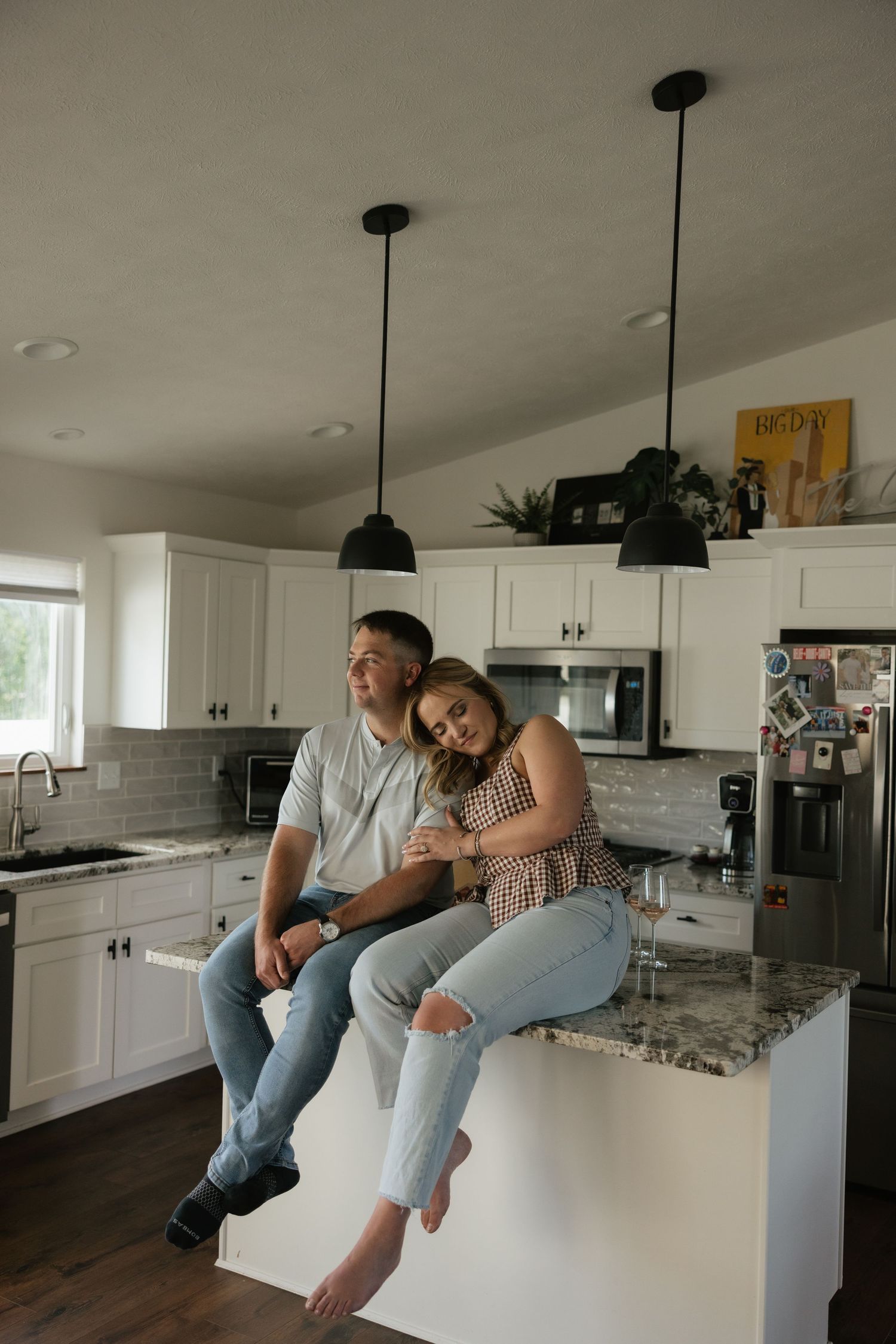 Couple sitting on kitchen island in modern white kitchen with pendant lights and granite countertops.