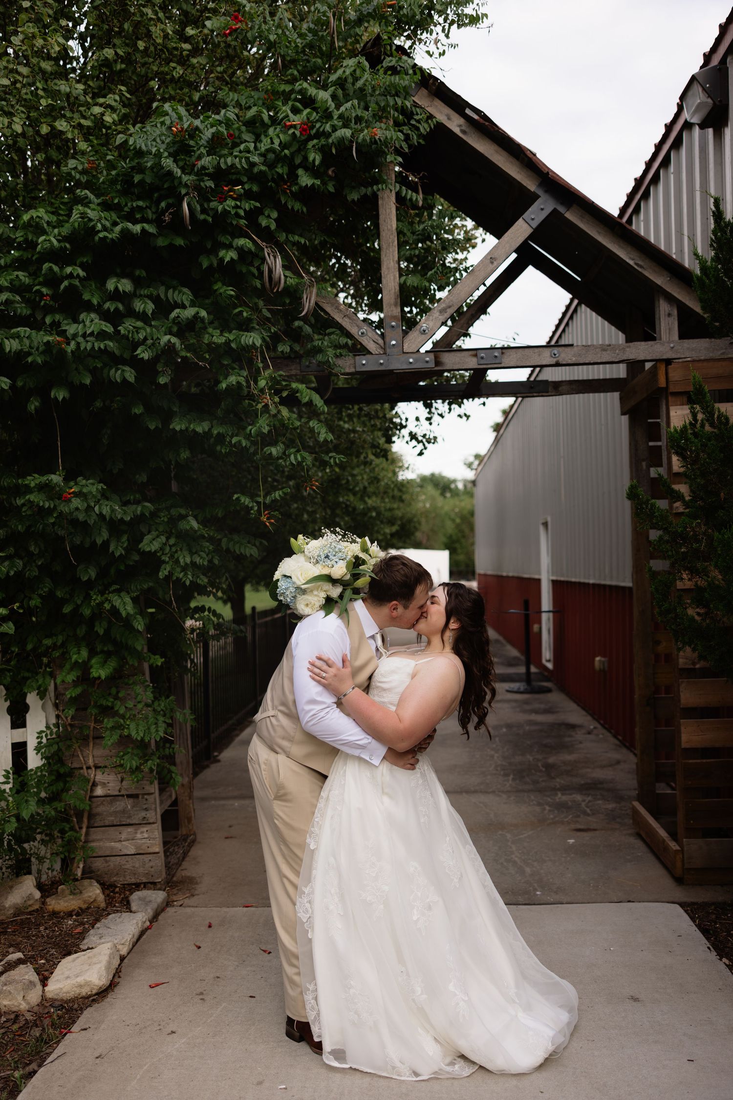 Bride and groom share a romantic kiss outdoors near a rustic barn, bride holding white bouquet in elegant wedding dress.