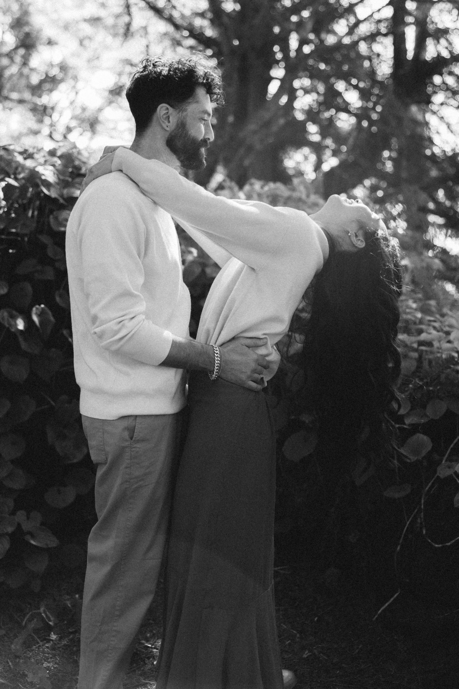 A couple embraces in black and white photography among garden foliage.