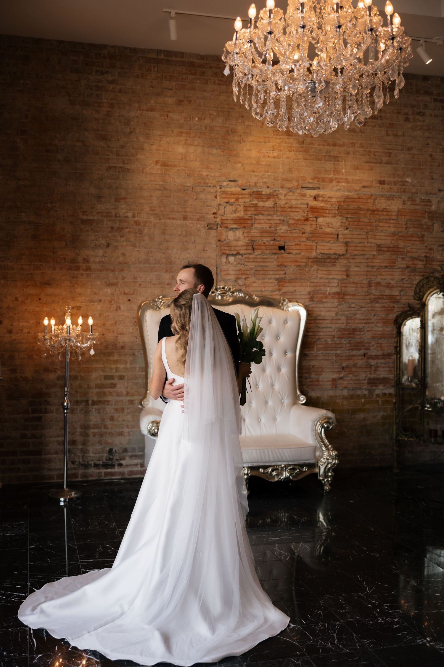 Bride and groom embrace in elegant venue with crystal chandelier, brick wall, and ornate white throne chair.