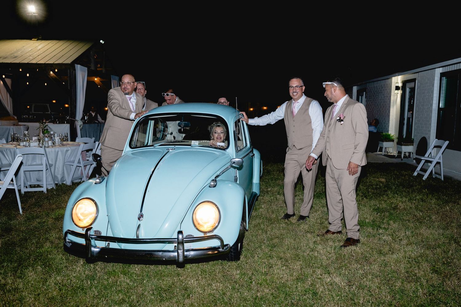 Men in suits pose with a vintage light blue VW Beetle car at an outdoor evening wedding reception.