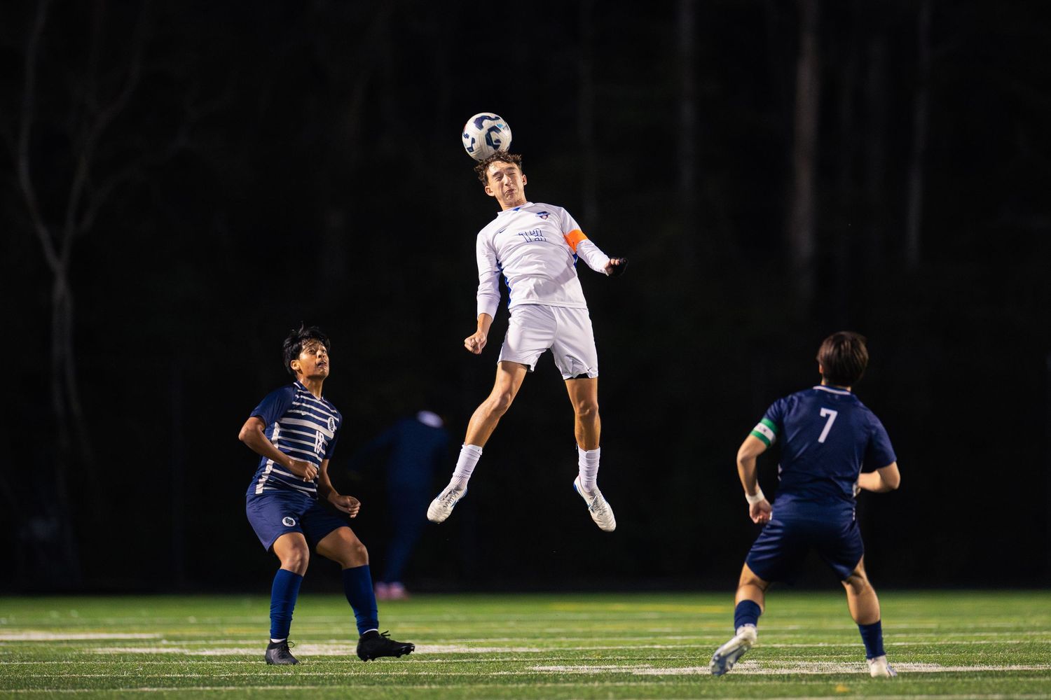 Soccer player leaps to head the ball during a night match on a lit field.