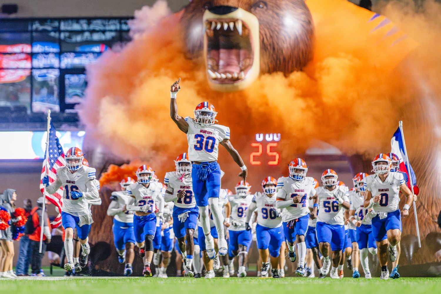 Grand Oaks High School football team runs onto field through dramatic orange smoke with roaring bear mascot.