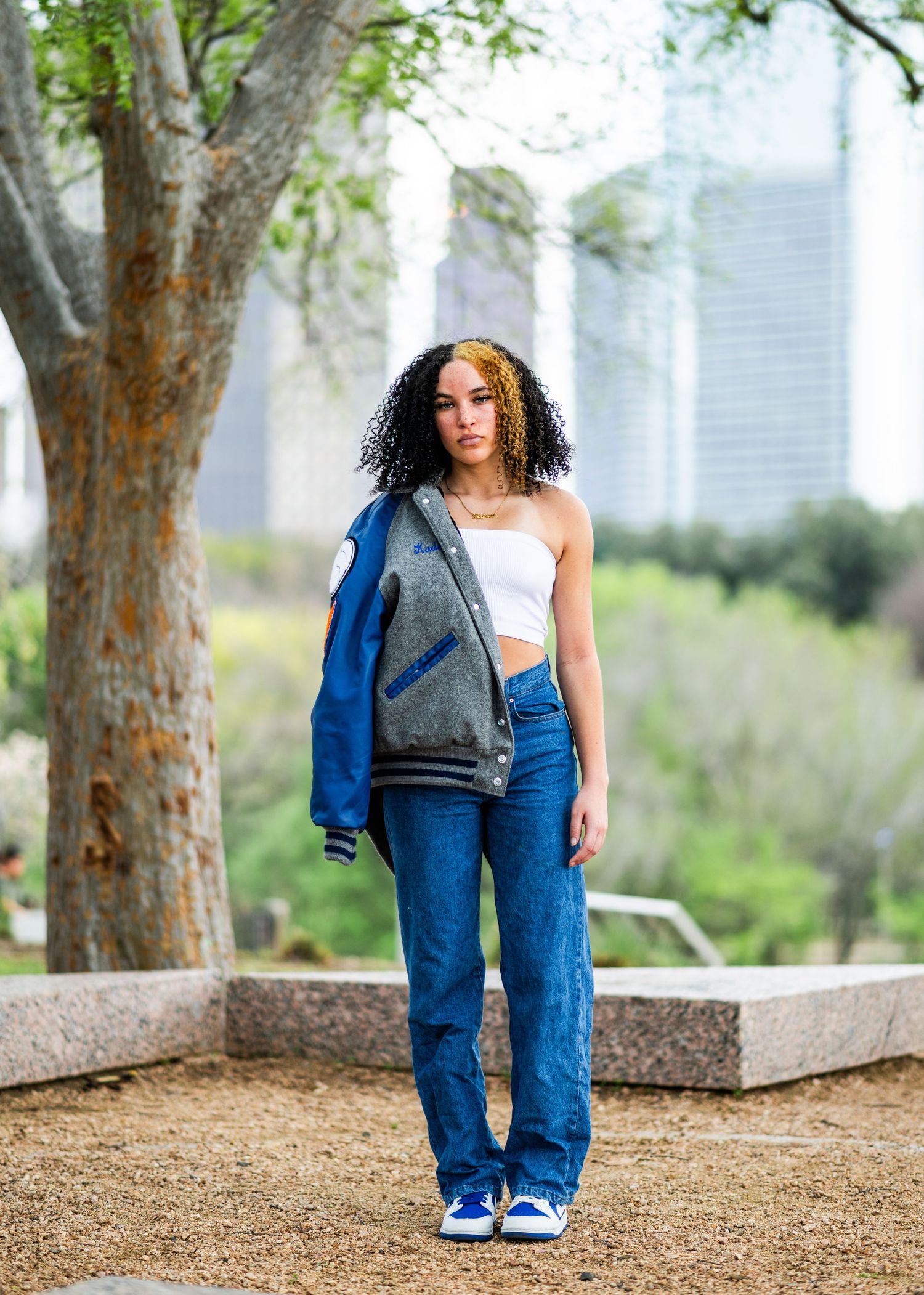 Young woman in varsity jacket and wide-leg jeans posing in urban park with skyline.