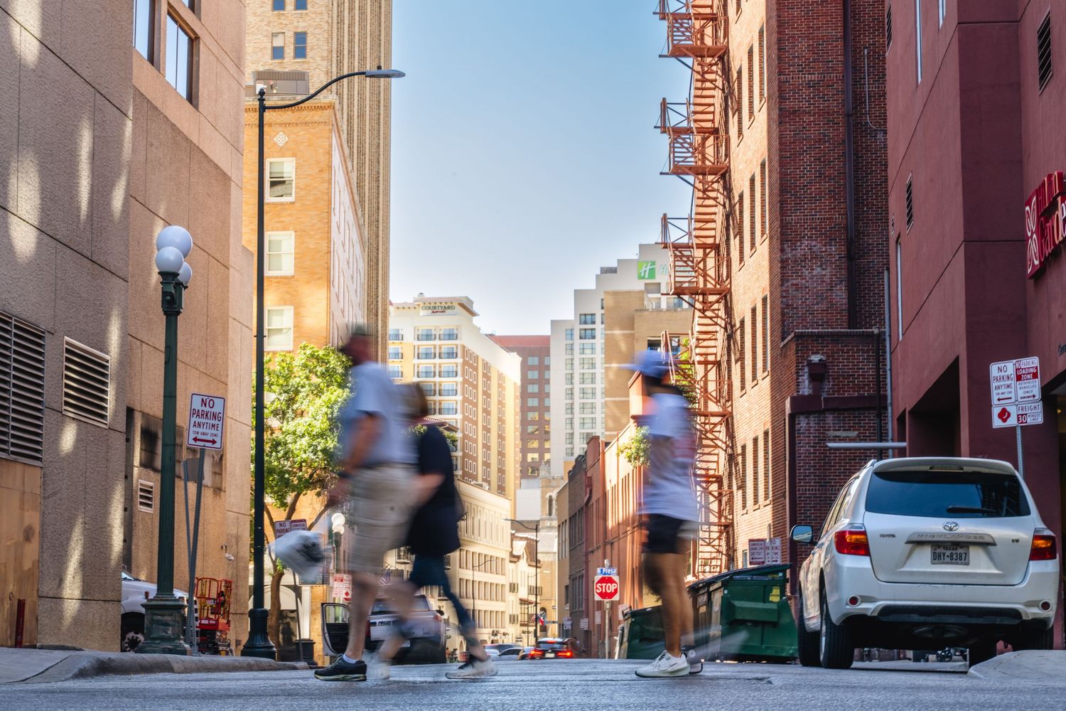 Blurred pedestrians and runners crossing a sunny urban street flanked by brick buildings.