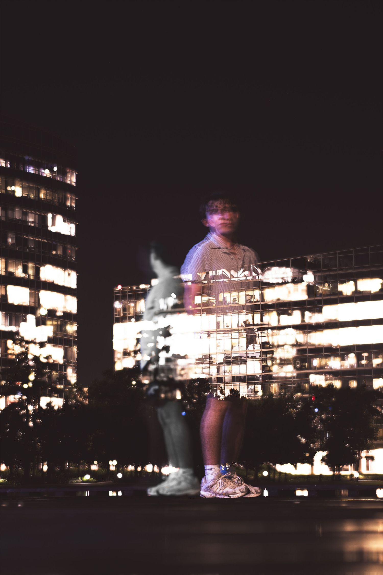 A long exposure night photo of a person standing in front of illuminated city buildings creating a ghostly blur effect.