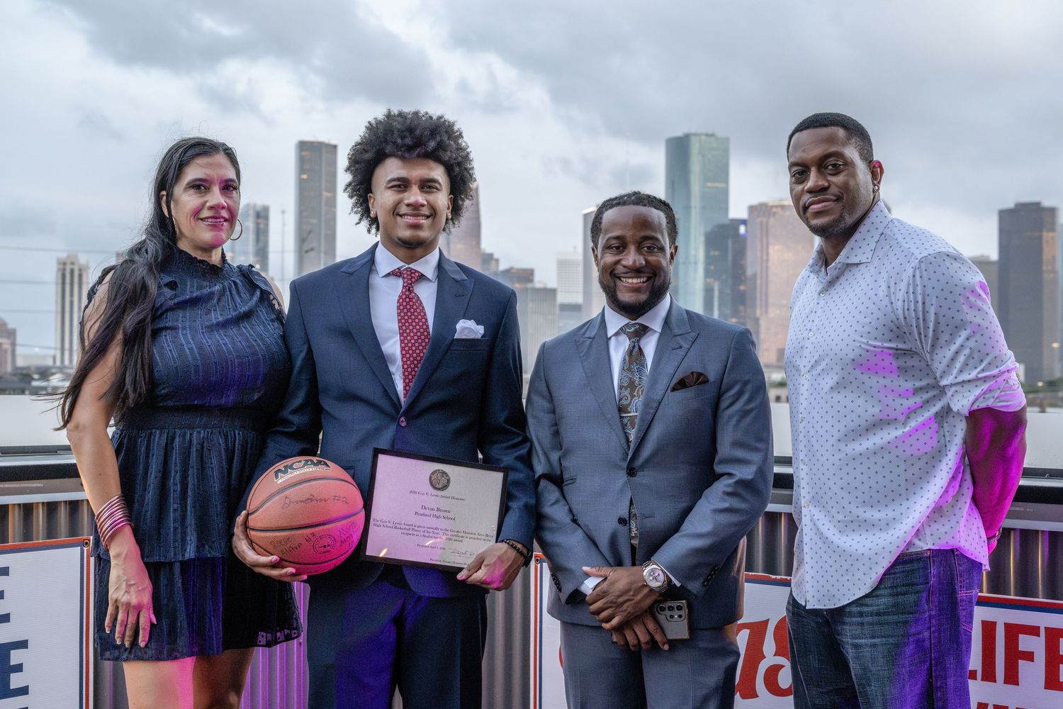 Four people pose on a rooftop with city skyline, young man holding basketball and certificate at awards ceremony.