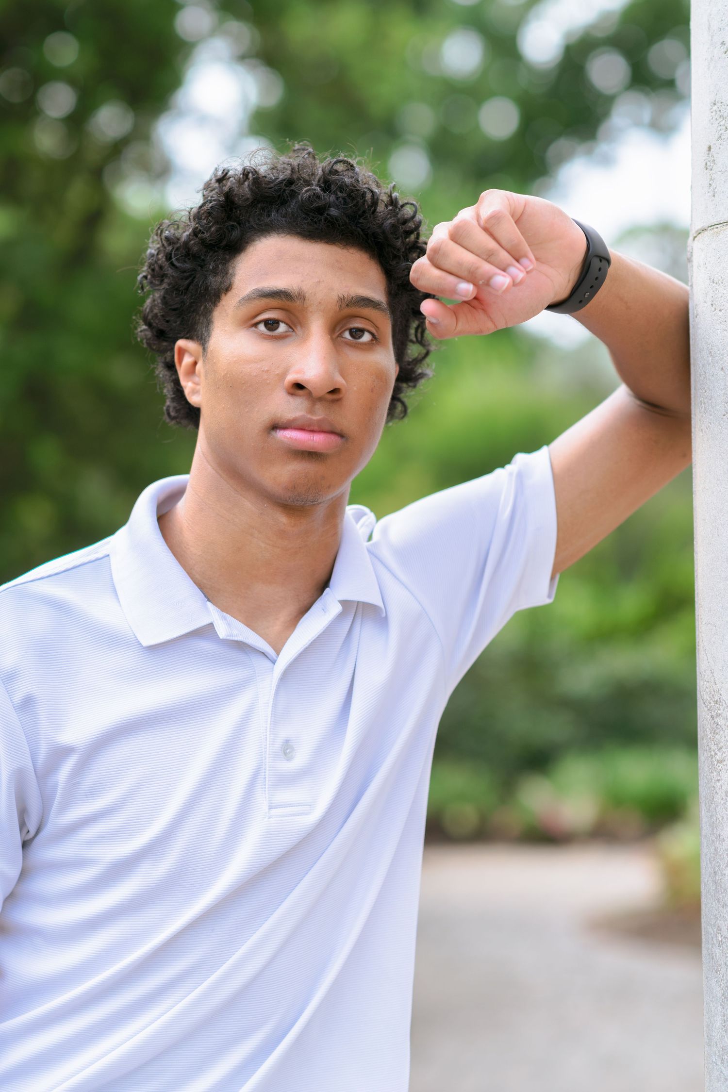 Young man with curly hair in white polo shirt leaning against pillar outdoors in green park setting.