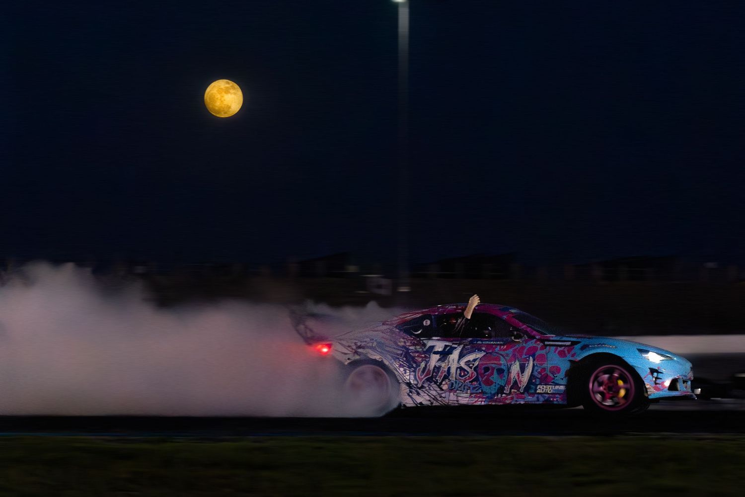 Colorful drift car performing a smoky drift under a full moon at night on a racing circuit.