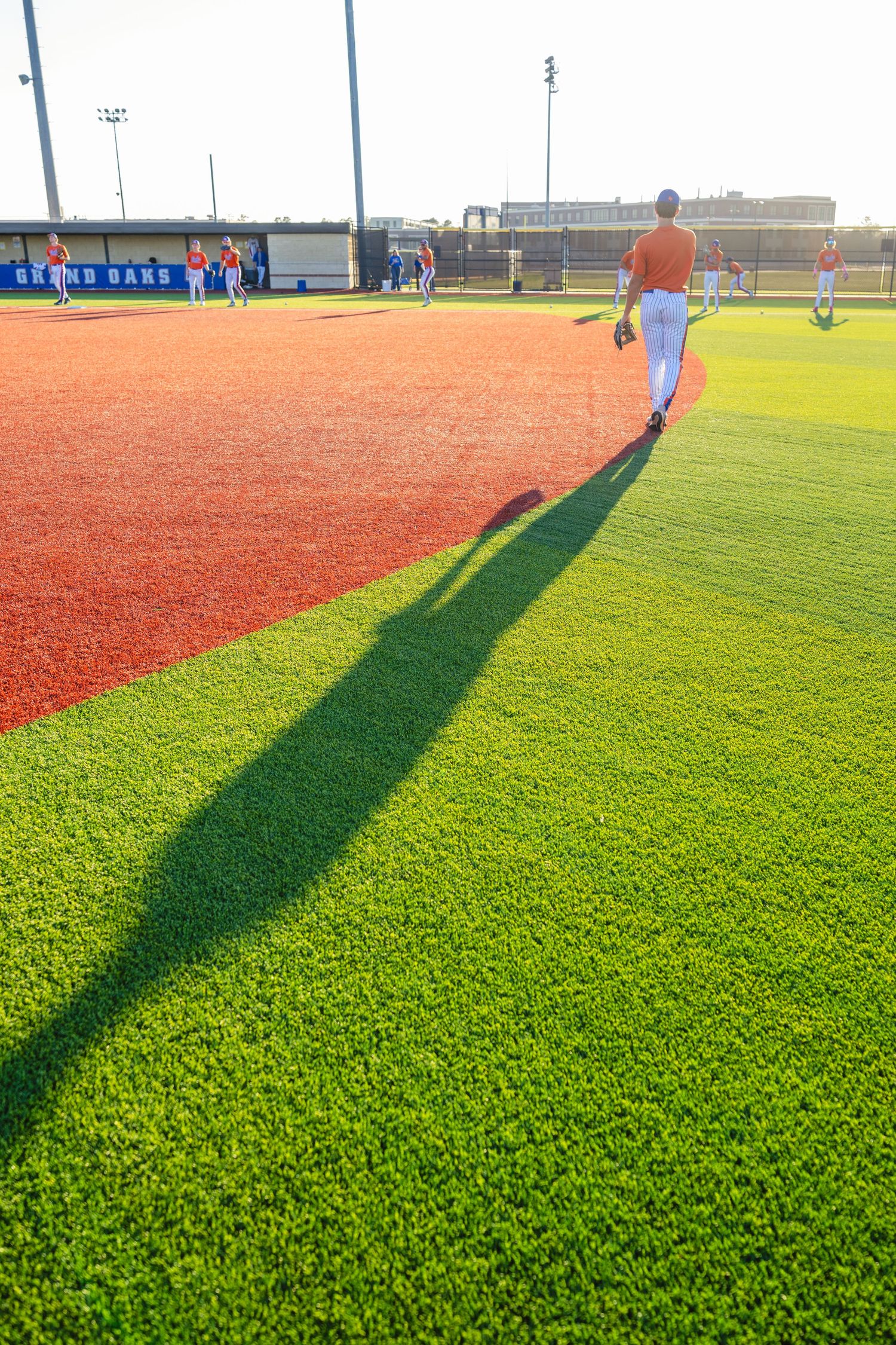 Baseball player walks toward red clay infield on bright green turf during sunny practice session.