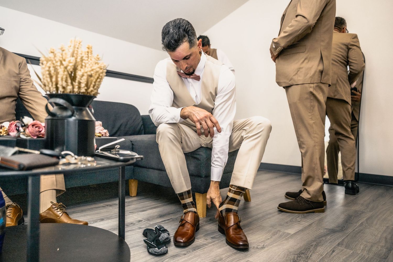 Groom putting on brown dress shoes while getting ready with groomsmen in suits in a modern room.
