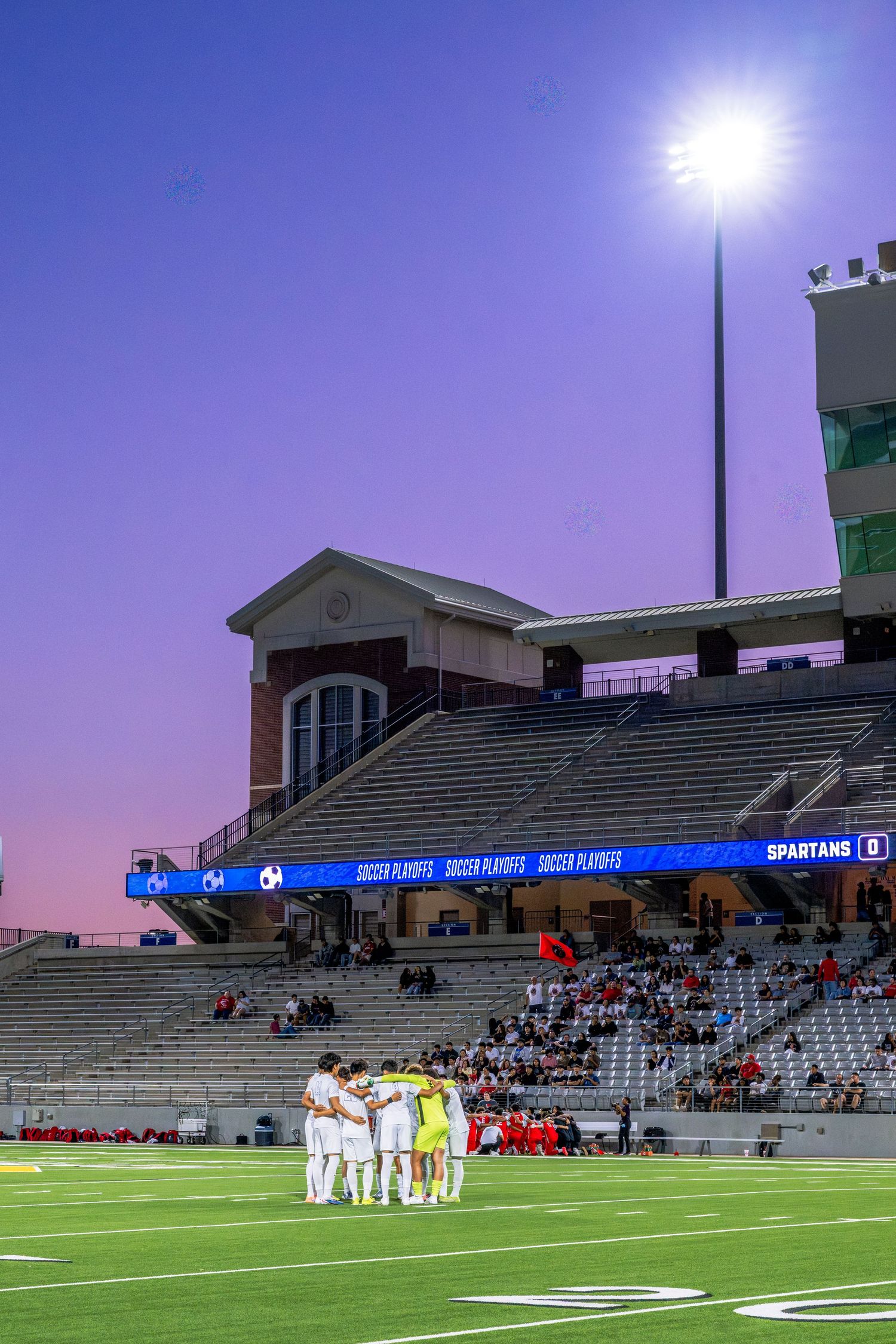 Soccer team huddles on field at dusk during playoff game at Spartans stadium.