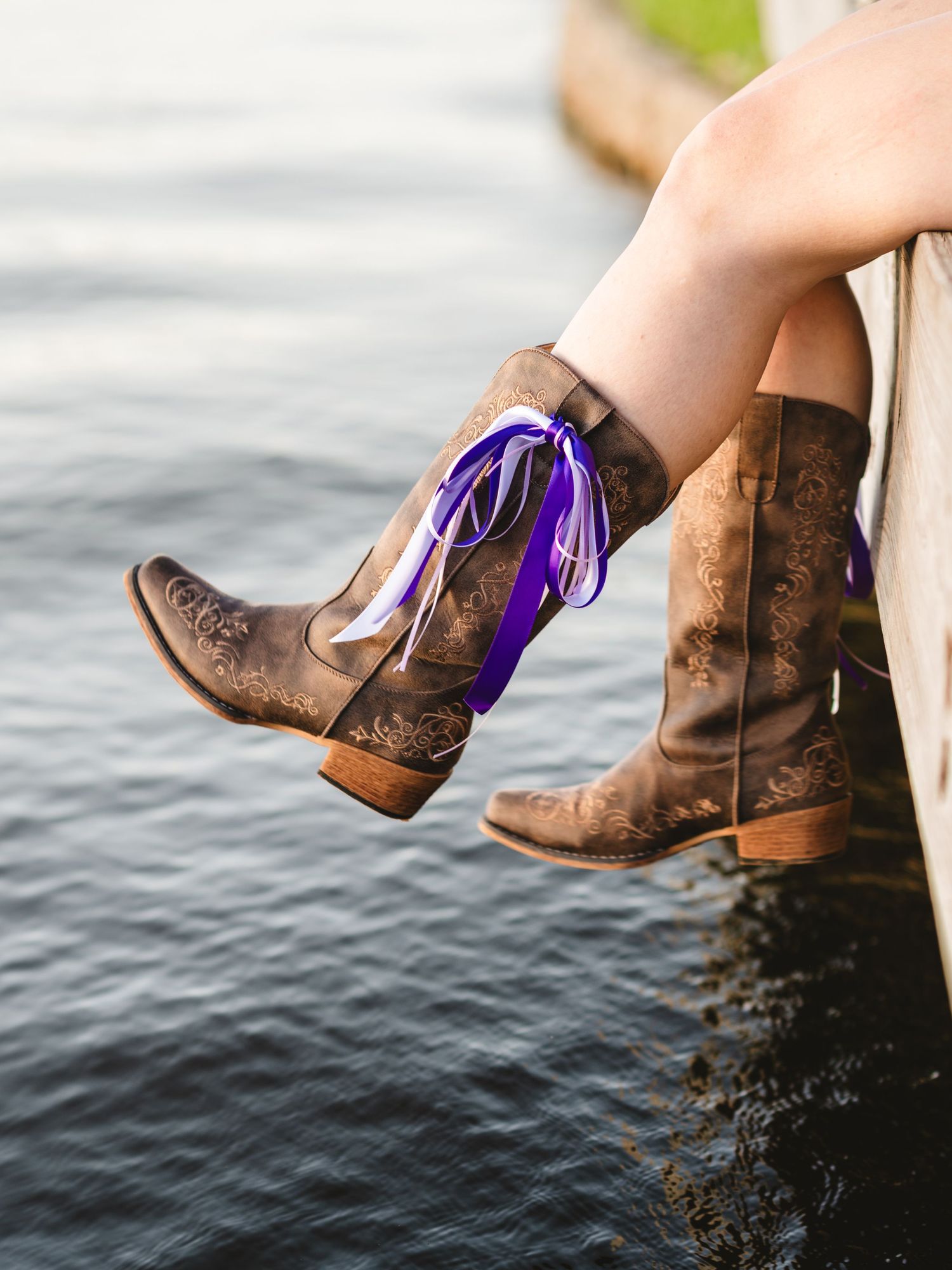 Close-up of brown cowboy boots with purple ribbon laces dangling over water's edge.