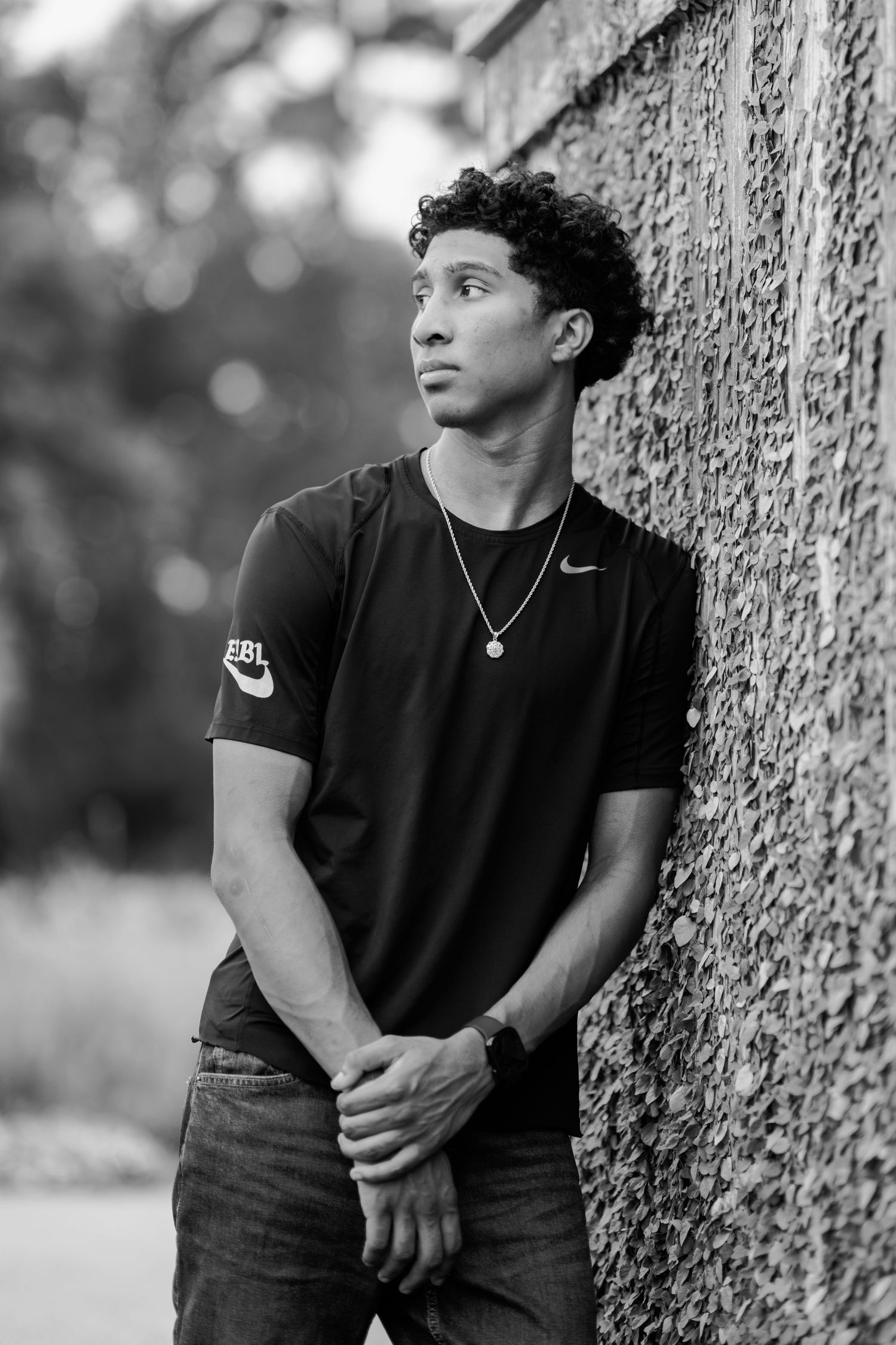 Black and white portrait of a young man in a Nike t-shirt leaning against a stone wall, looking thoughtfully upward.